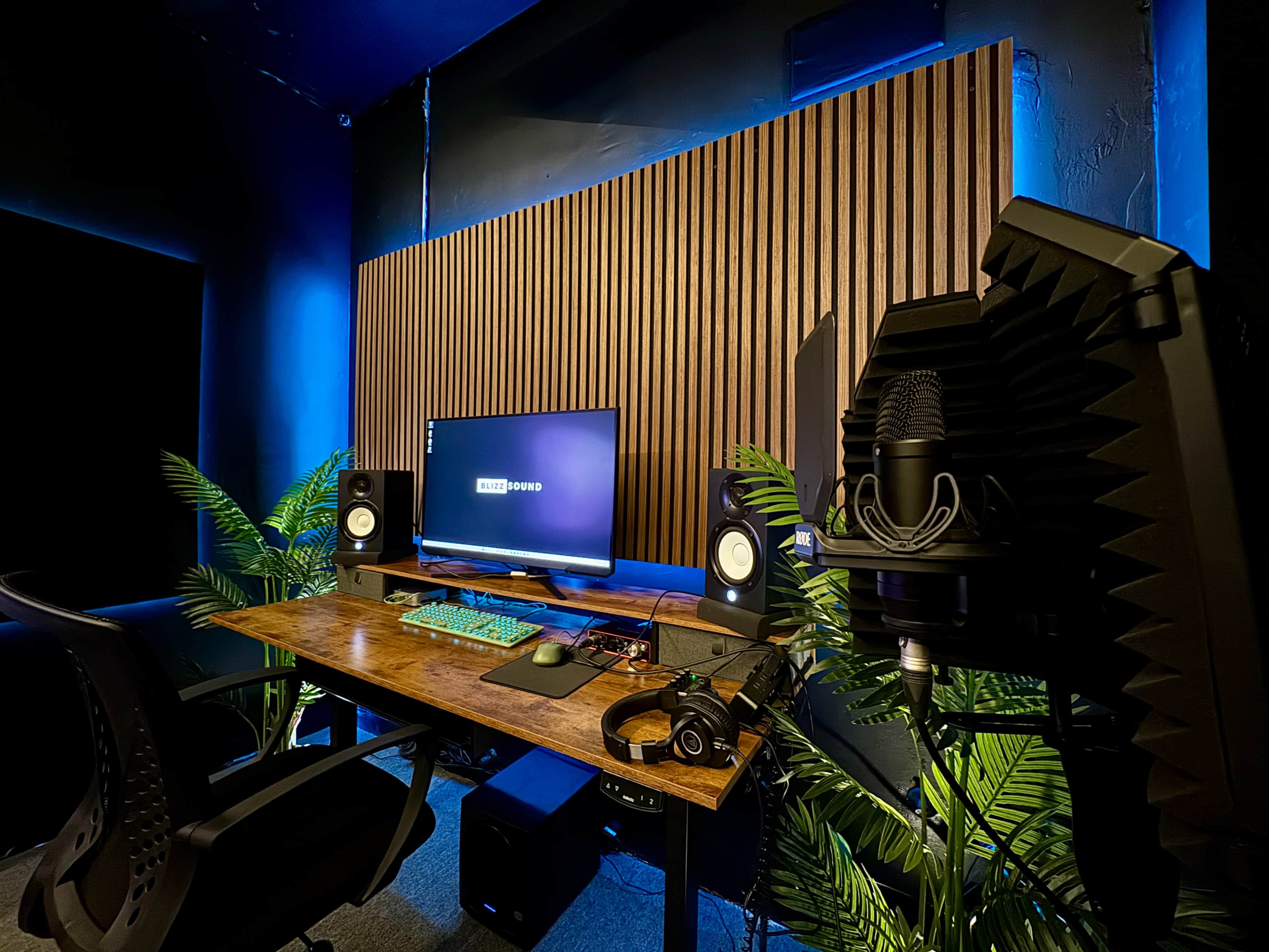 The image shows a modern home office setup featuring a wooden desktop with a computer monitor, speakers, a microphone, headphones, and decorative plants against a backdrop of a paneled wall illuminated by blue light.