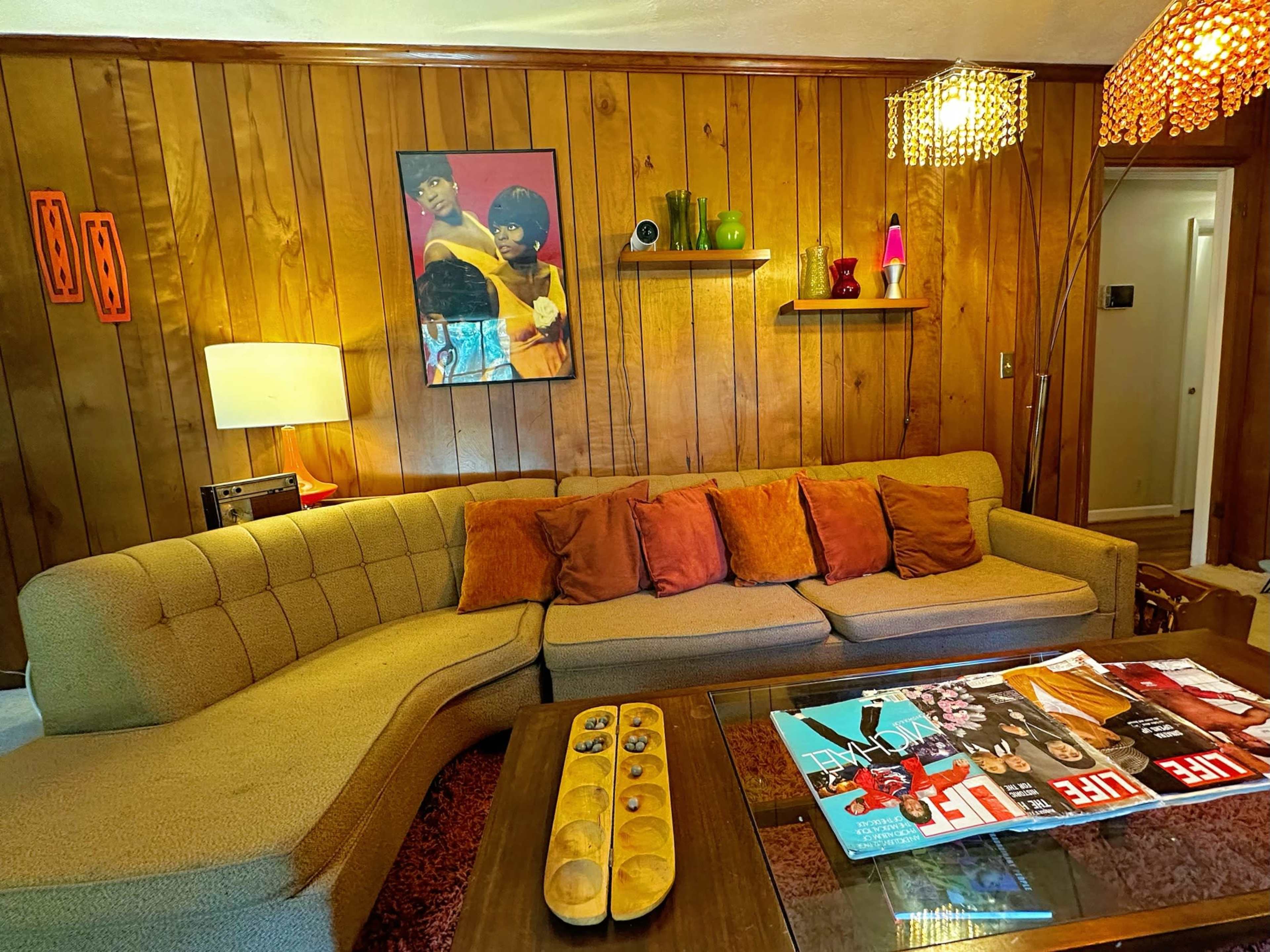 The image shows a cozy living room featuring a curved sofa with orange pillows, a wooden wall, and various decorative items on shelves and a coffee table.
