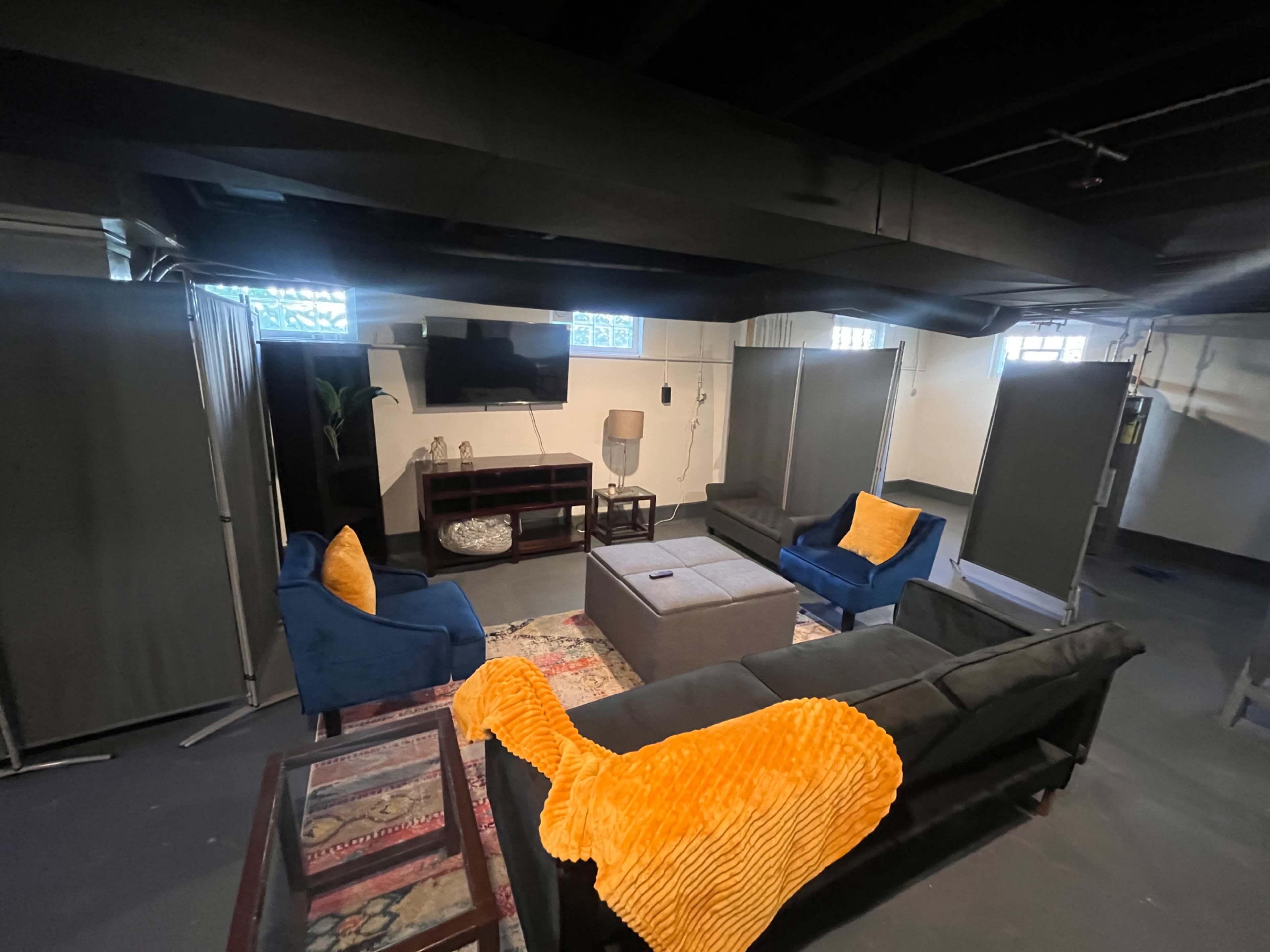 The image shows a dimly lit basement lounge area with two blue sofas, a gray coffee table, and various partitions creating a cozy seating arrangement.