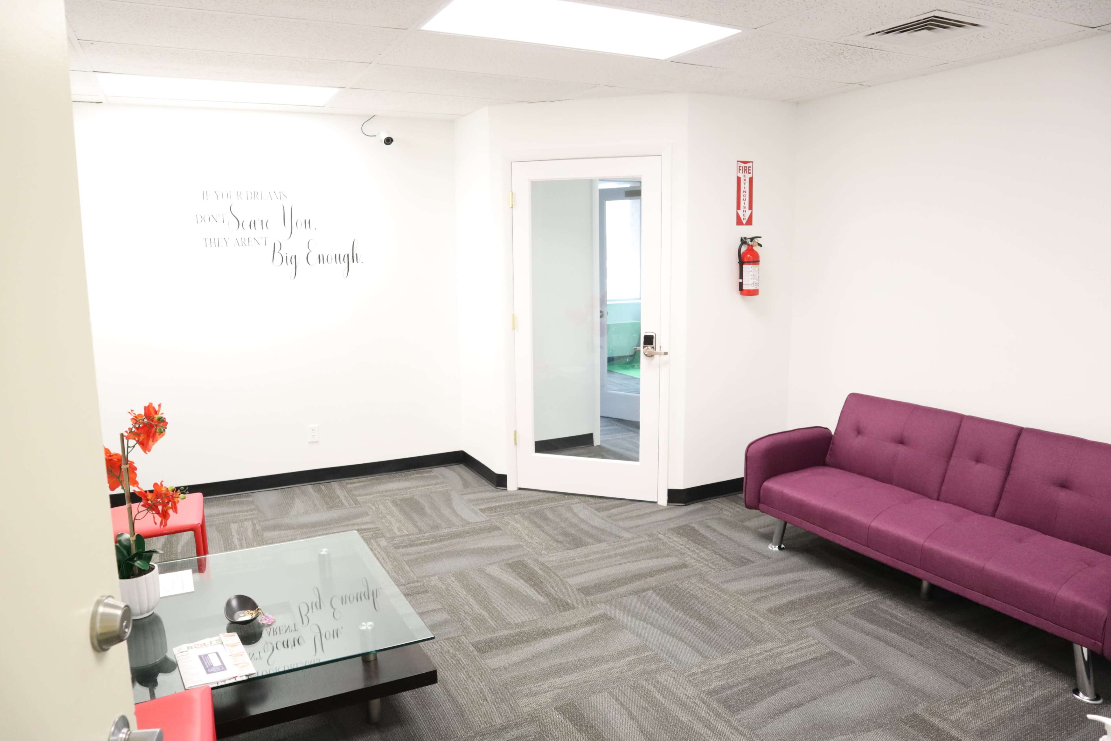 A waiting area features a purple couch, a glass coffee table, and a door leading outside, with a wall displaying a motivational quote.