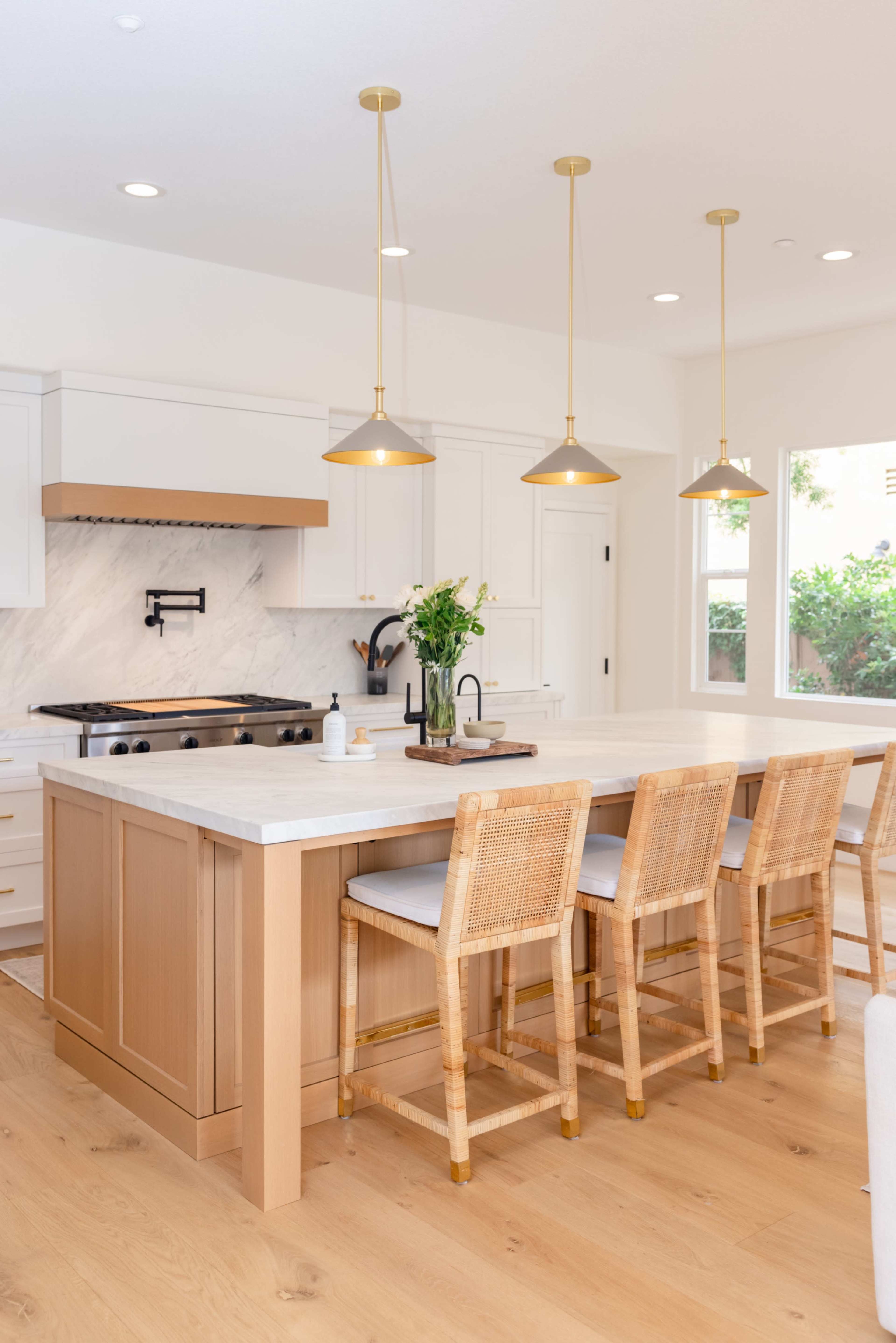 A modern kitchen features a large island with bar stools, gold pendant lights, and a marble backsplash behind the stove.