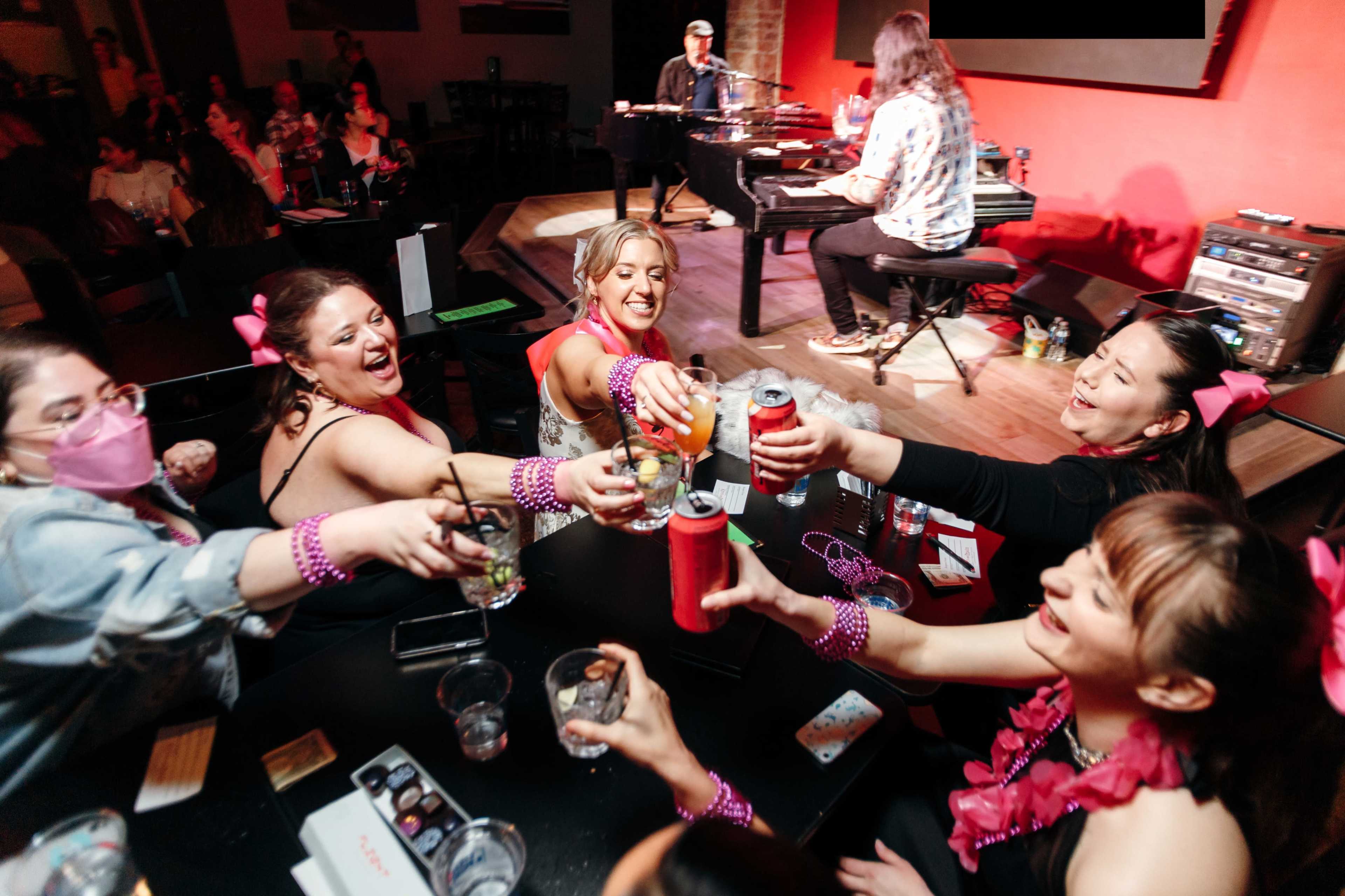 A group of women in festive attire raise their drinks in a lively bar while a musician plays a piano on stage.