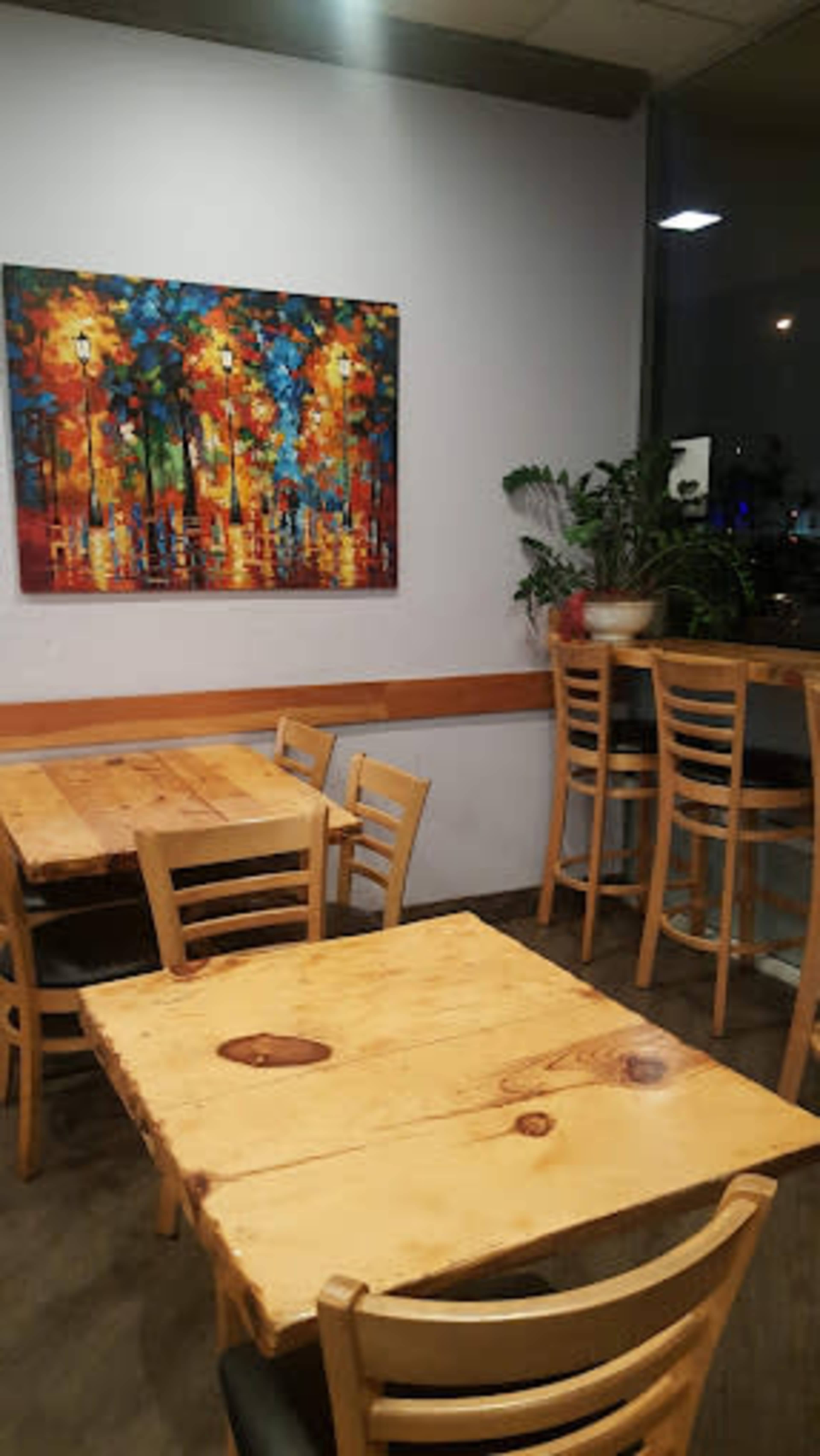 The image shows a cozy café interior featuring wooden tables and chairs, with a vibrant painting on the wall and a potted plant in the corner.