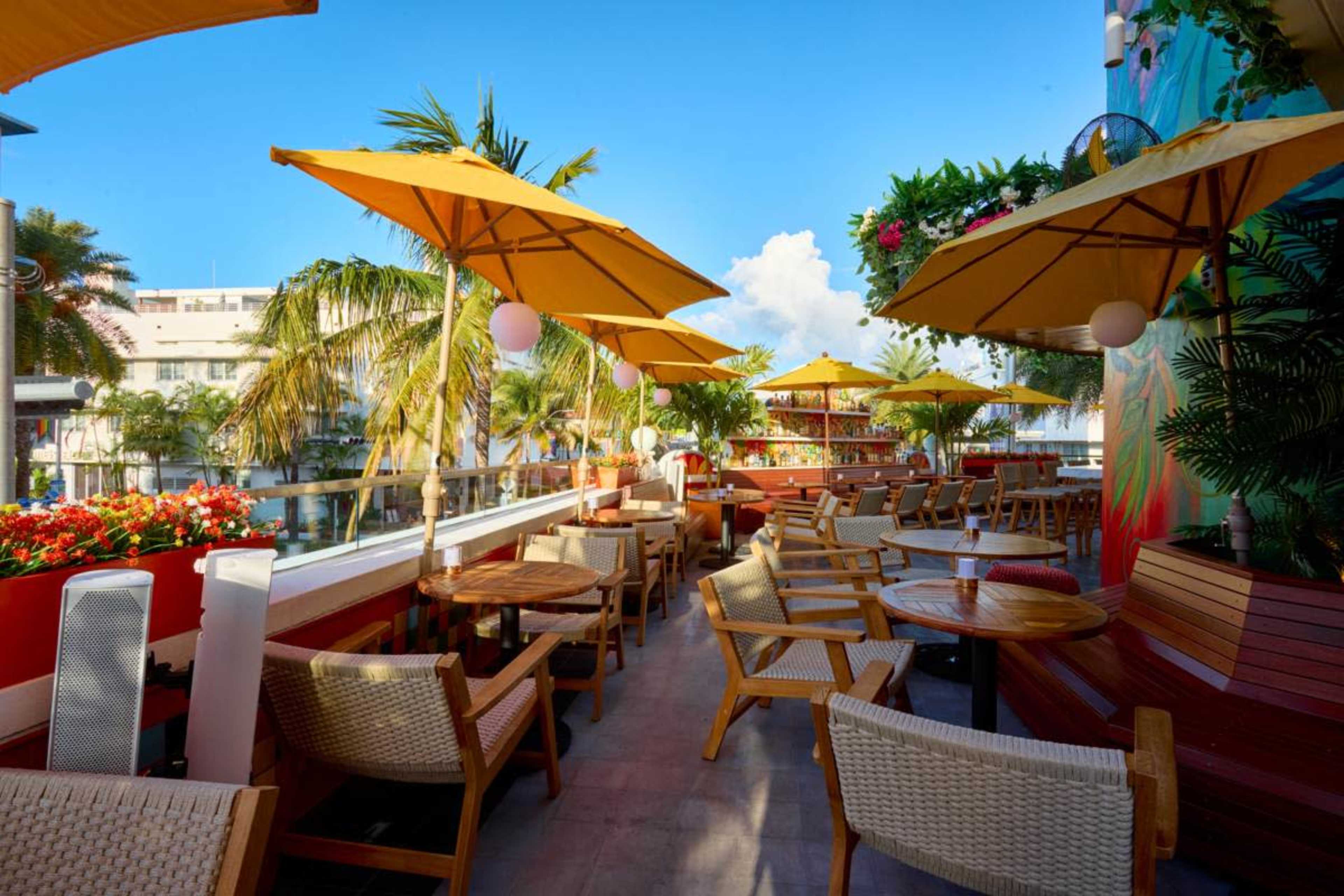 A vibrant outdoor restaurant patio features wooden tables and chairs, adorned with yellow umbrellas, surrounded by tropical plants and flowers.