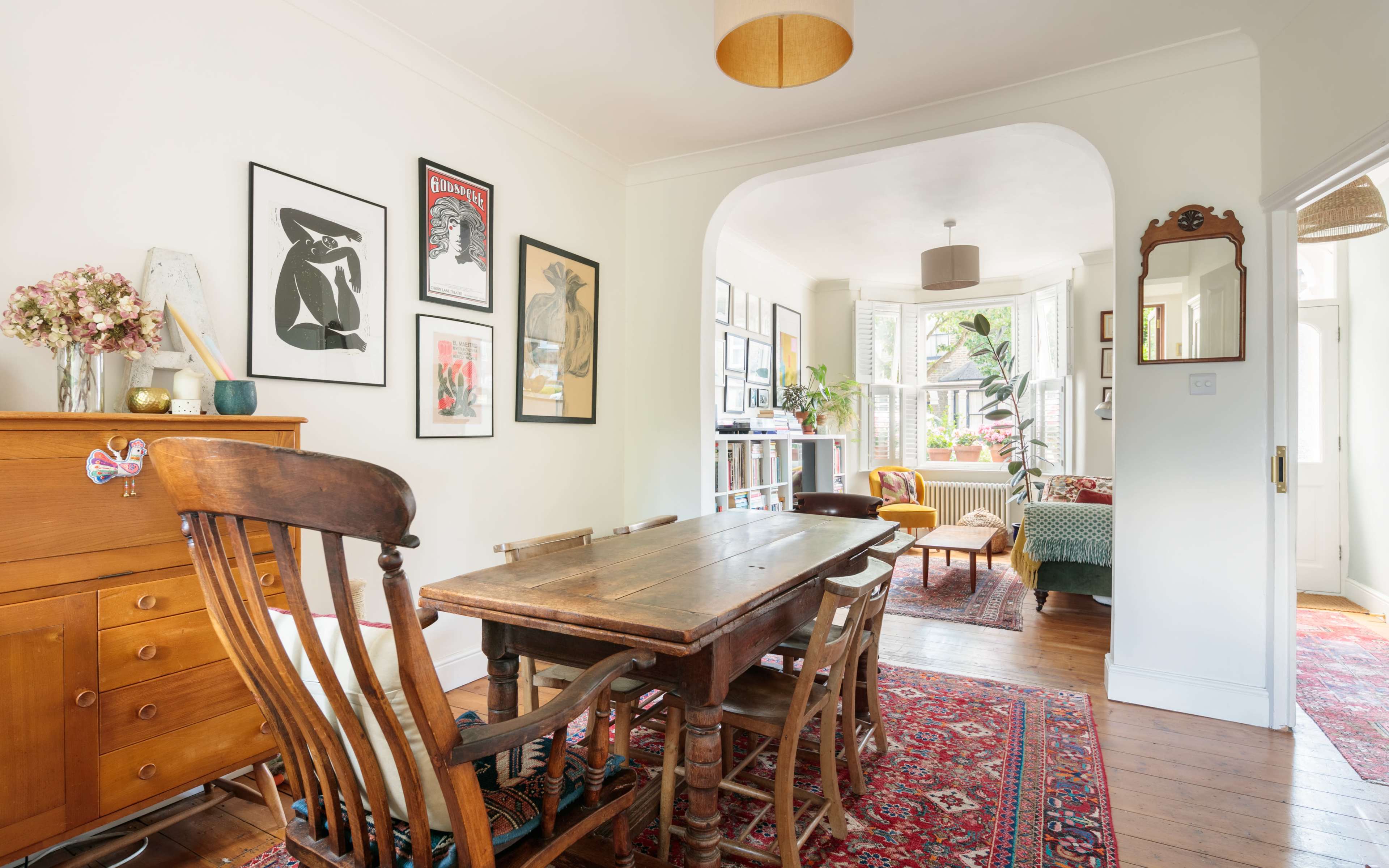 A bright dining area with a wooden table surrounded by chairs, framed artwork on the walls, and a view into a cozy, well-lit living space.