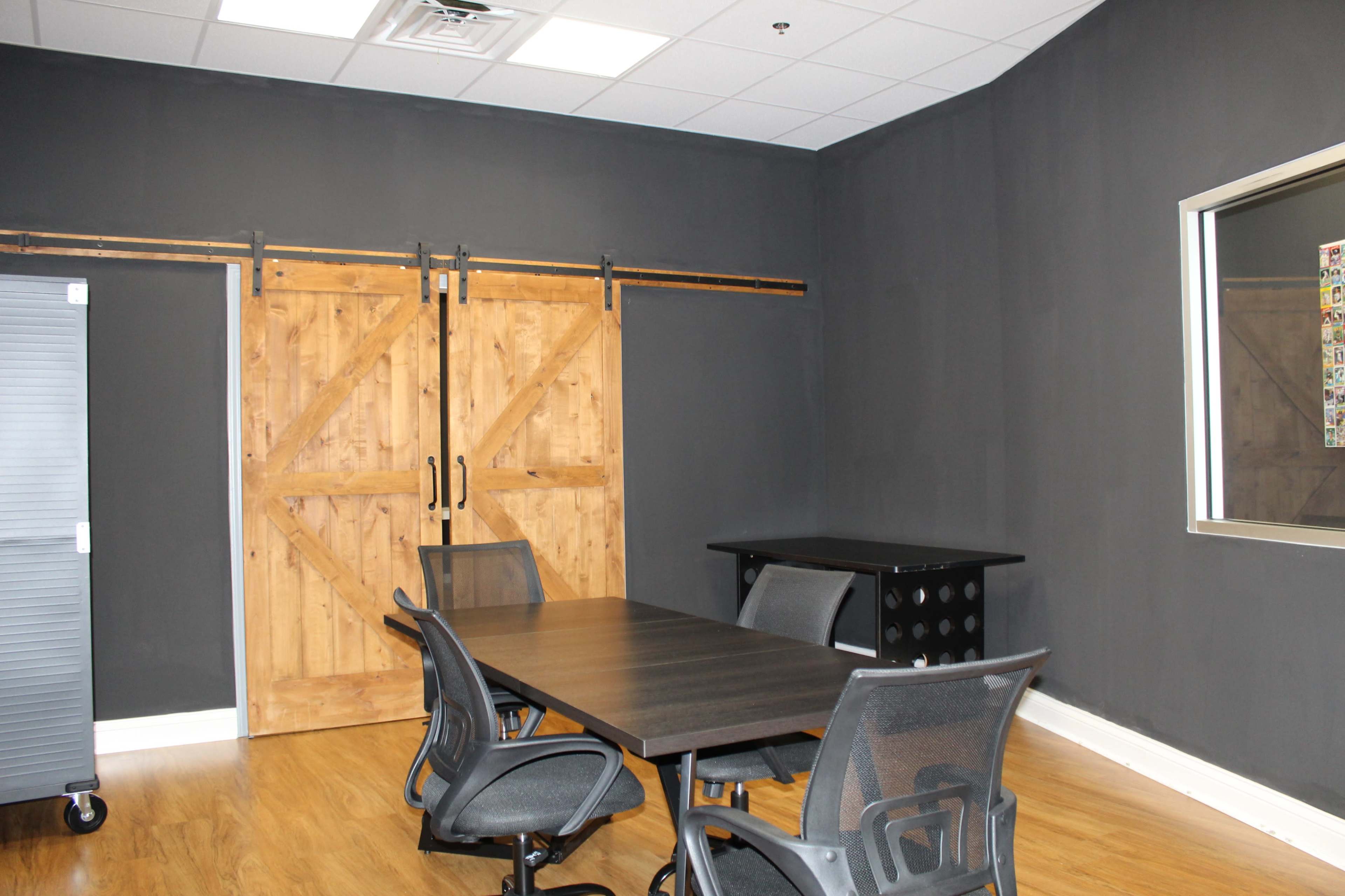 The image shows a conference room with a wooden sliding door, a black conference table, and several black office chairs against a dark wall.