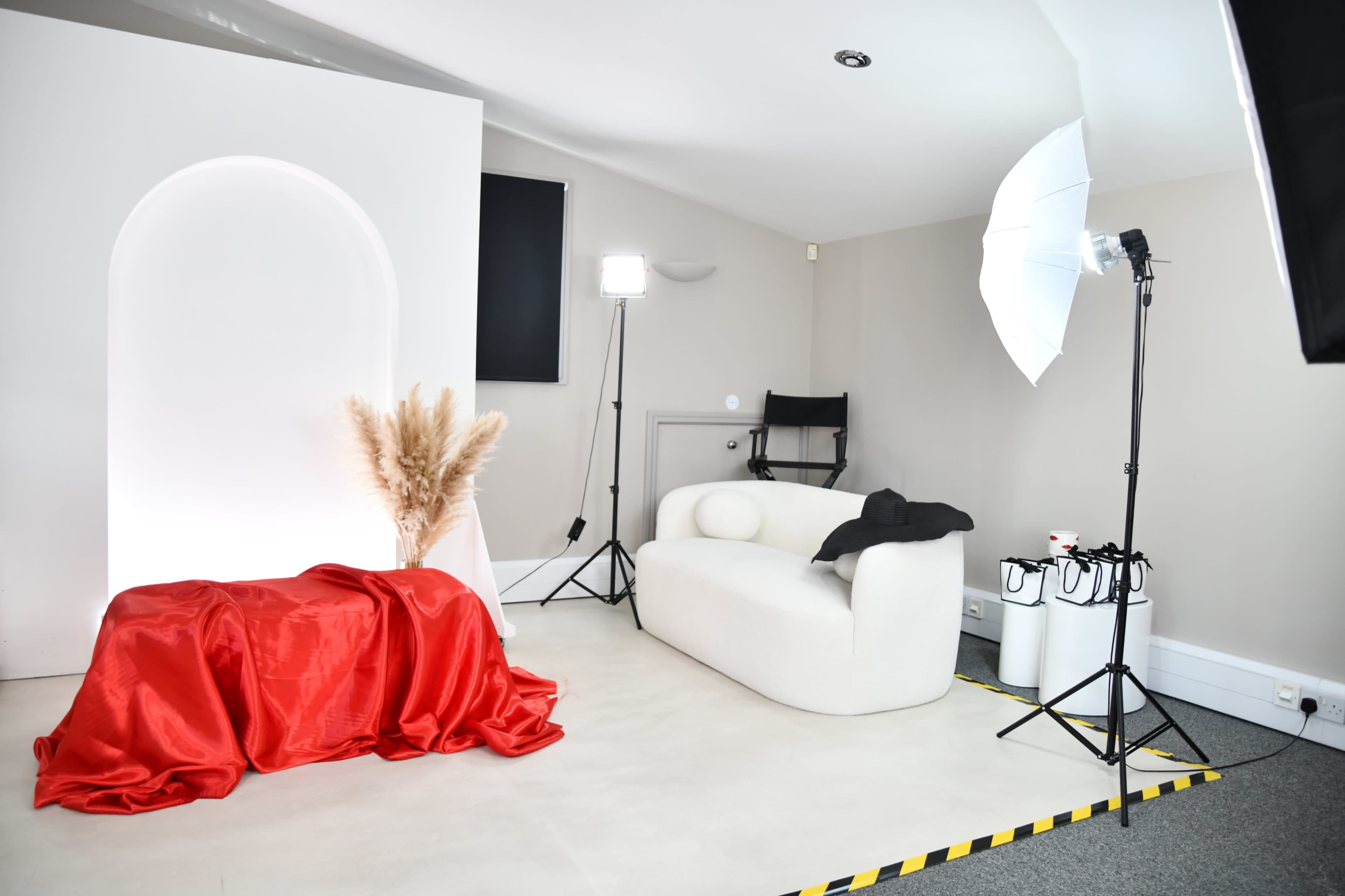 The image features a studio setup with a white backdrop, a red fabric draped over a table, a white modern couch, and lighting equipment arranged around the space.