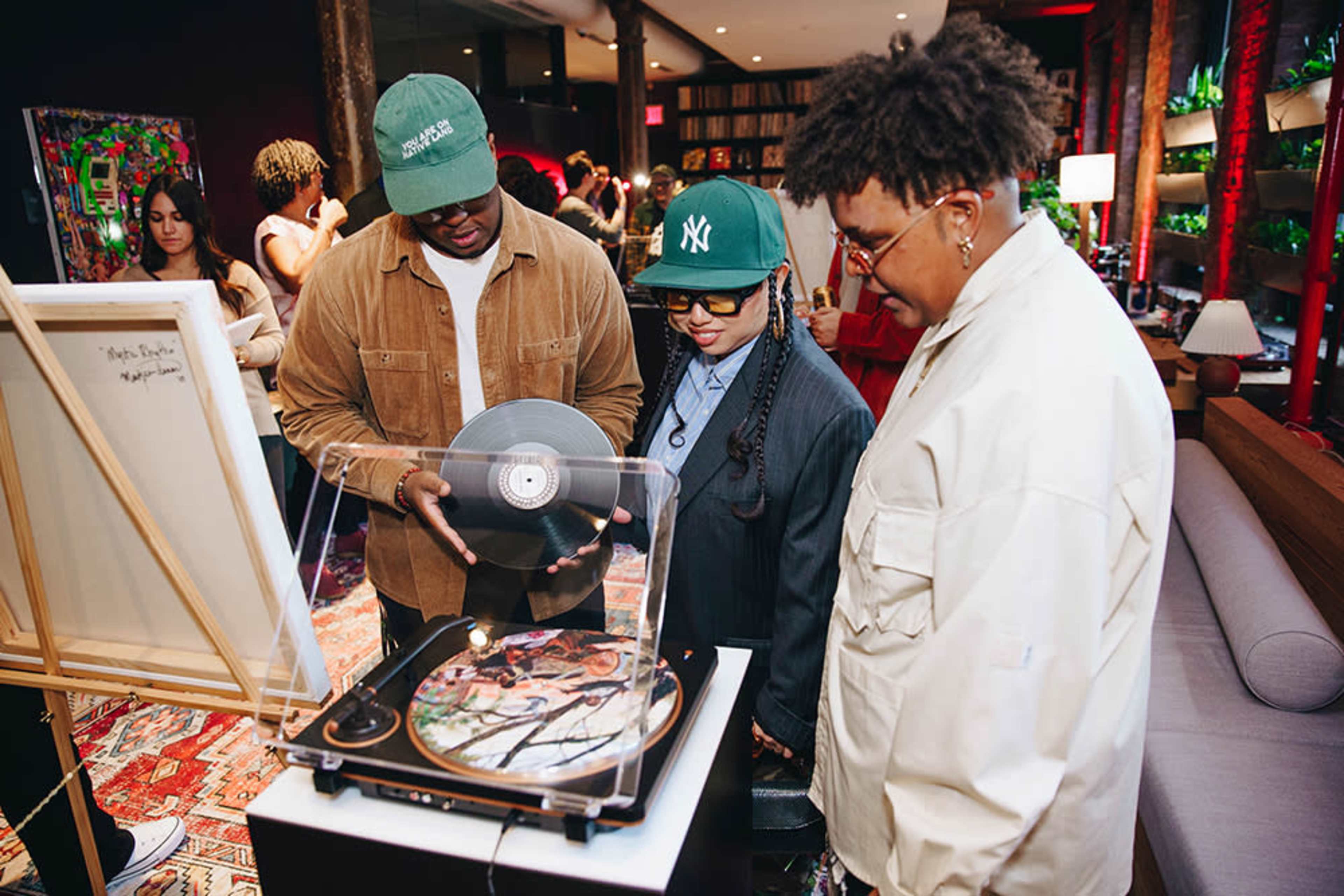 Three people are gathered around a record player, with one person holding a vinyl record while another looks on, in a stylish indoor space filled with books and art.
