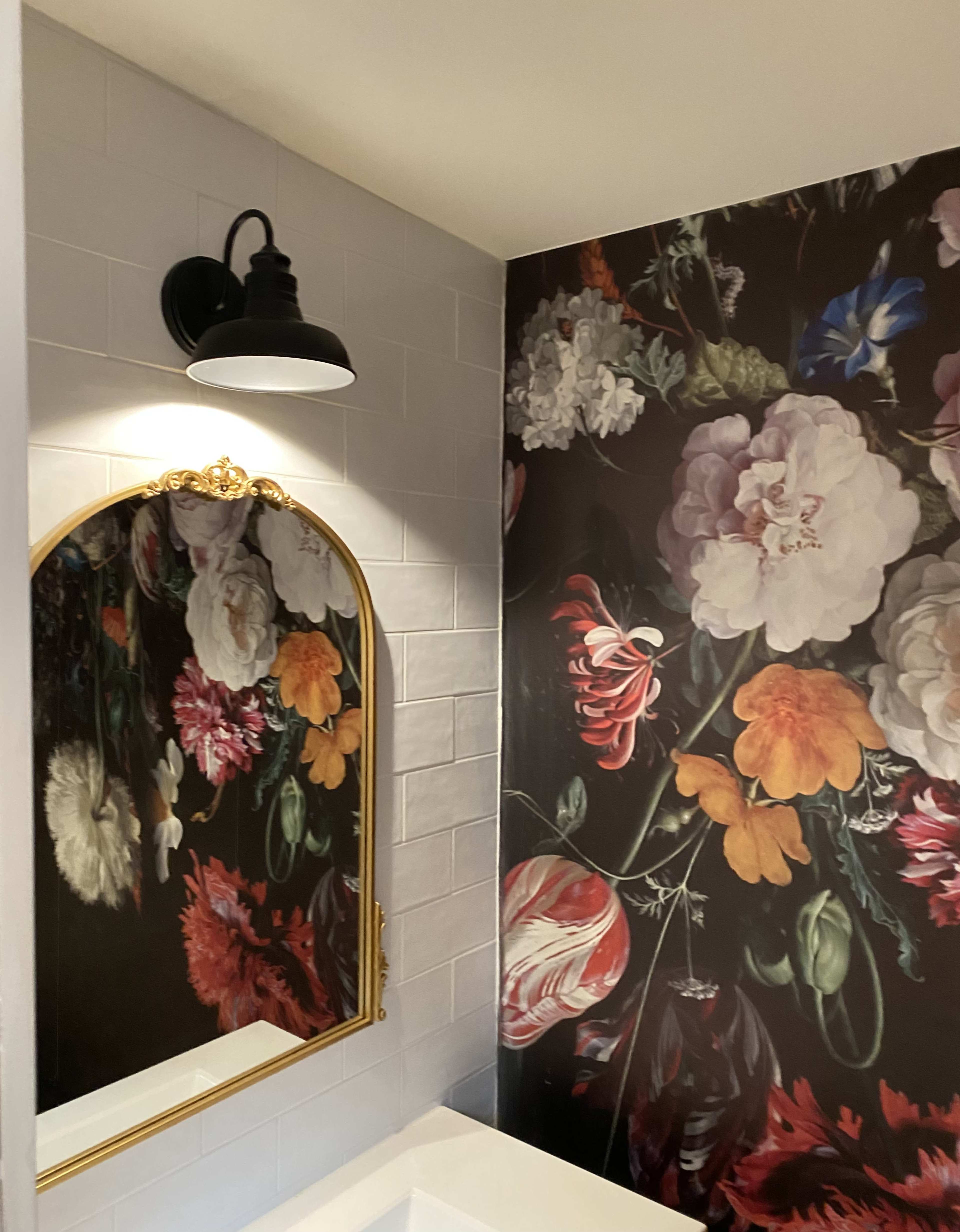 A bathroom corner featuring a large floral wallpaper design, a white tiled wall, and a golden-framed mirror illuminated by a wall-mounted light fixture.