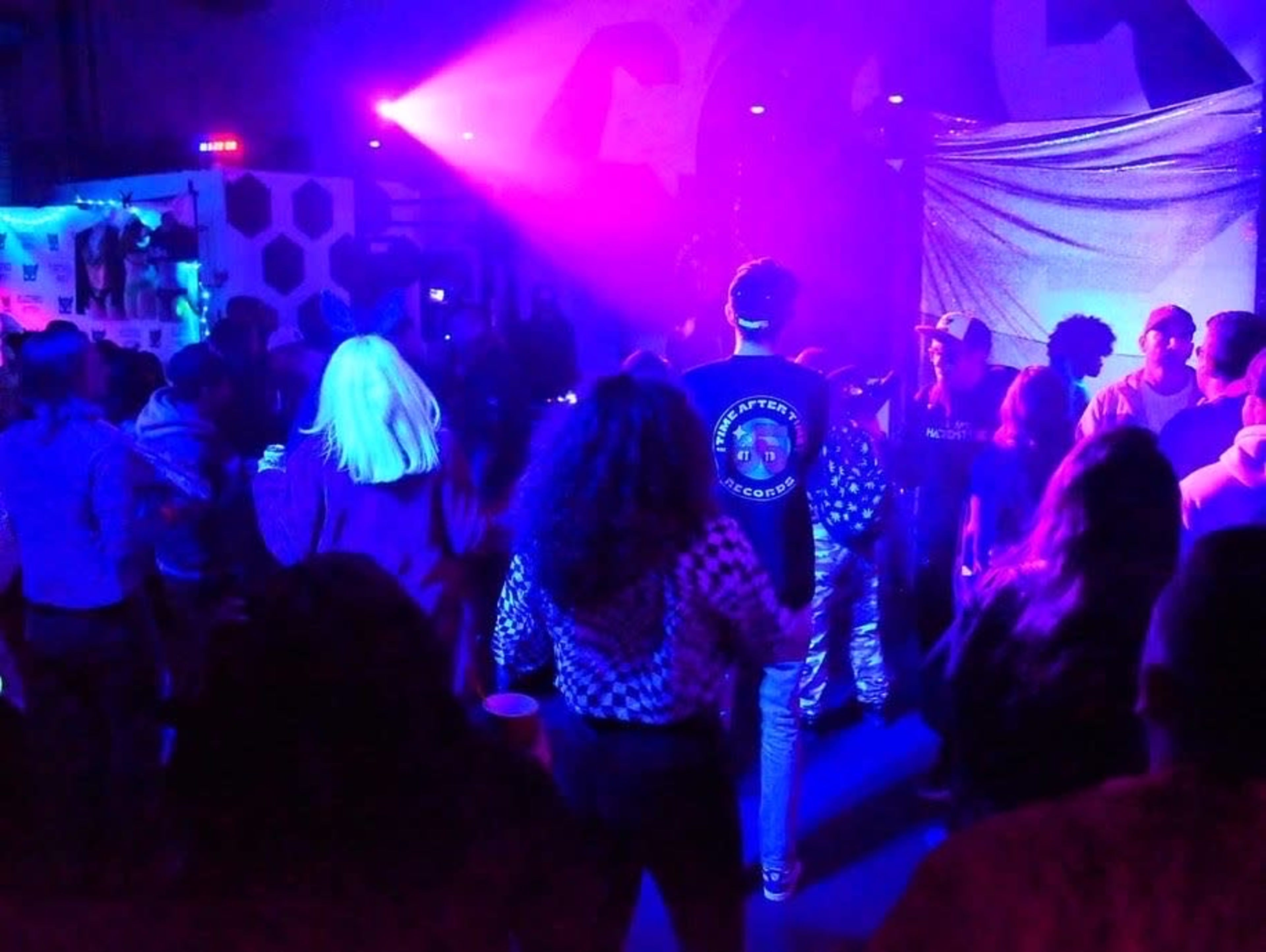 A crowd of people dances under purple and blue lights in a lively indoor setting.