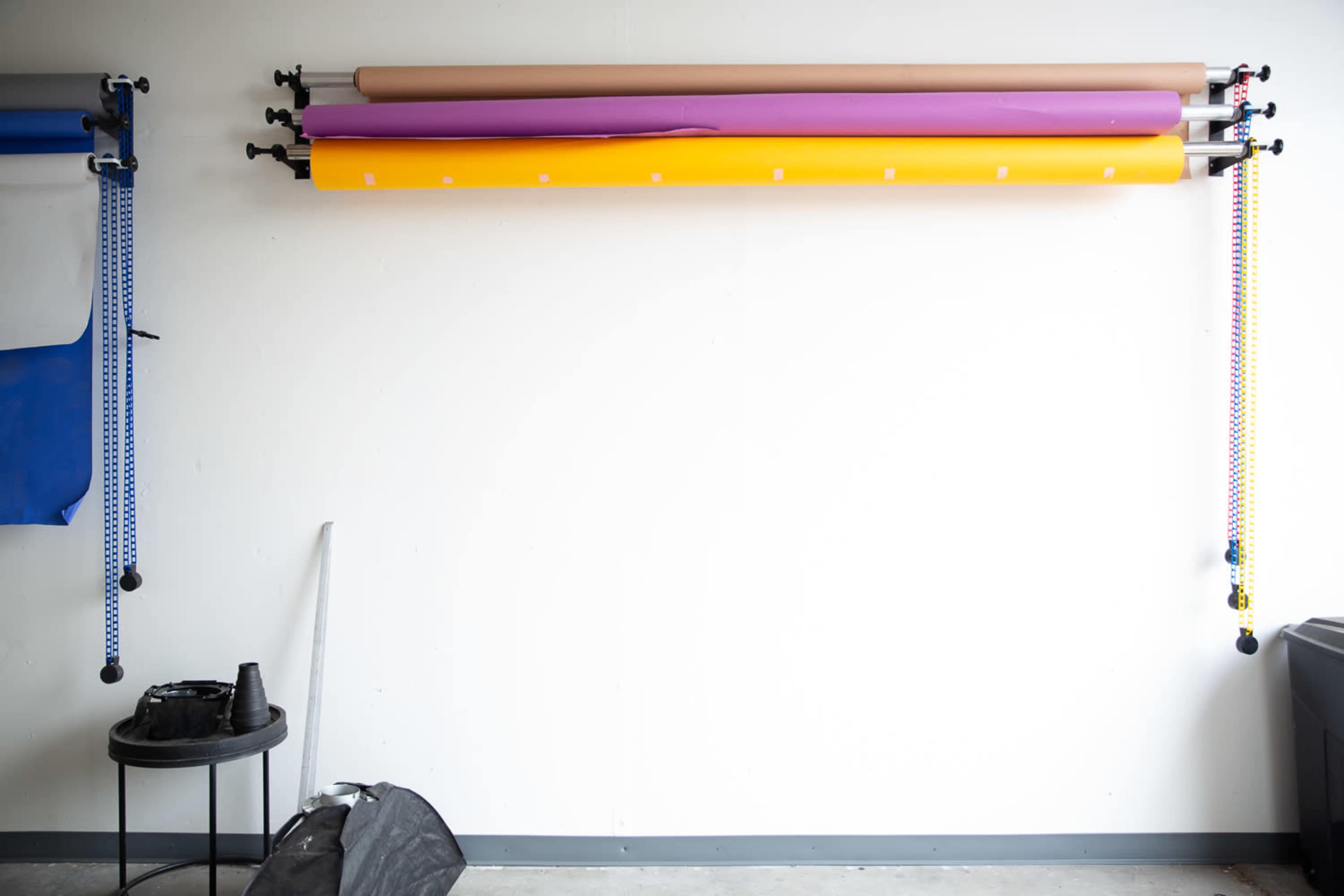 The image shows a wall with several rolls of colored backdrop paper mounted on a rack, alongside a black stool and equipment on the floor.
