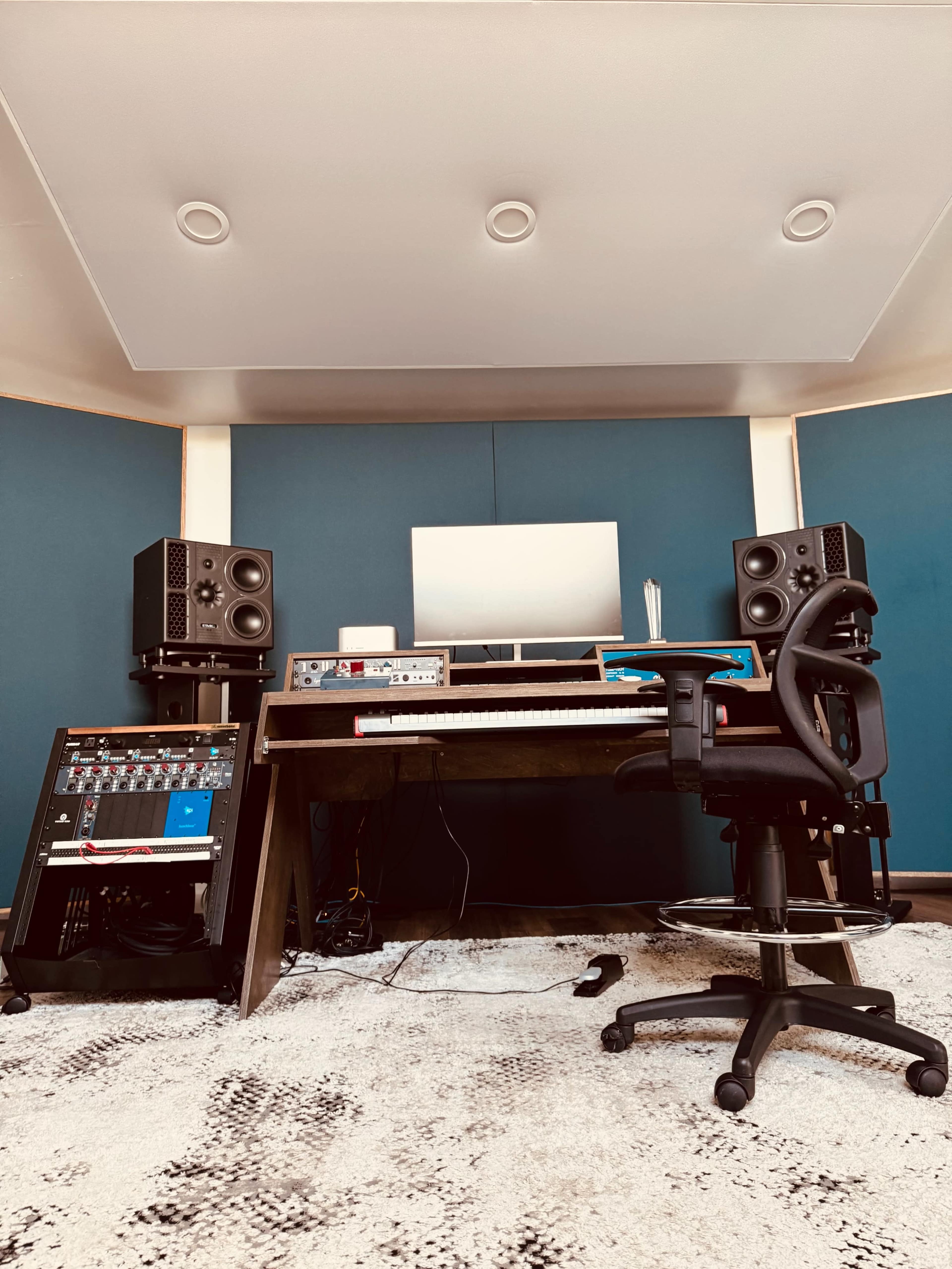 The image shows a music production studio with a desk supporting audio monitors, a computer, and various equipment, all set against blue acoustic panels.