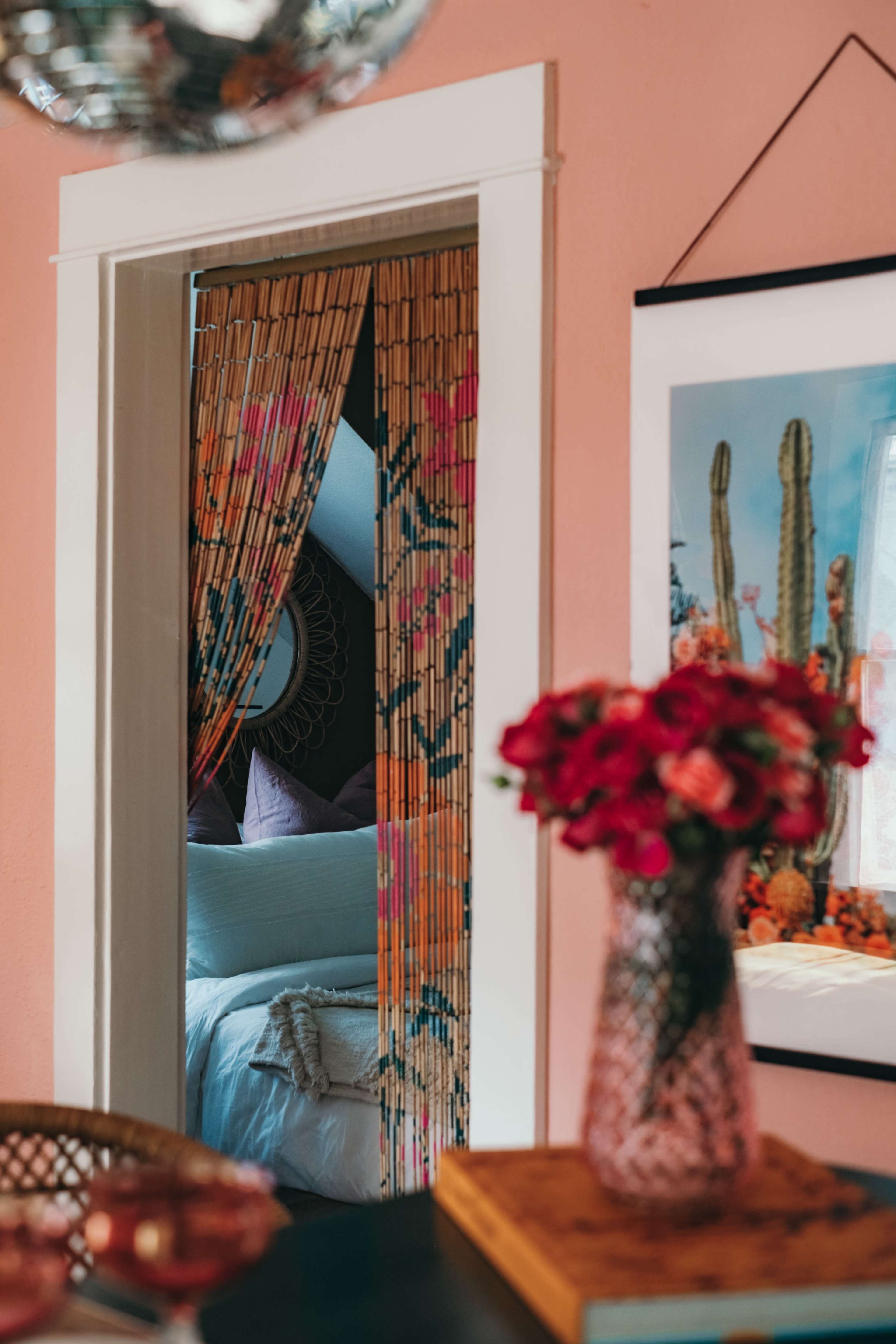 A doorway adorned with bamboo beads leads into a softly lit room, featuring a bed and decorative elements in a pink-walled space.