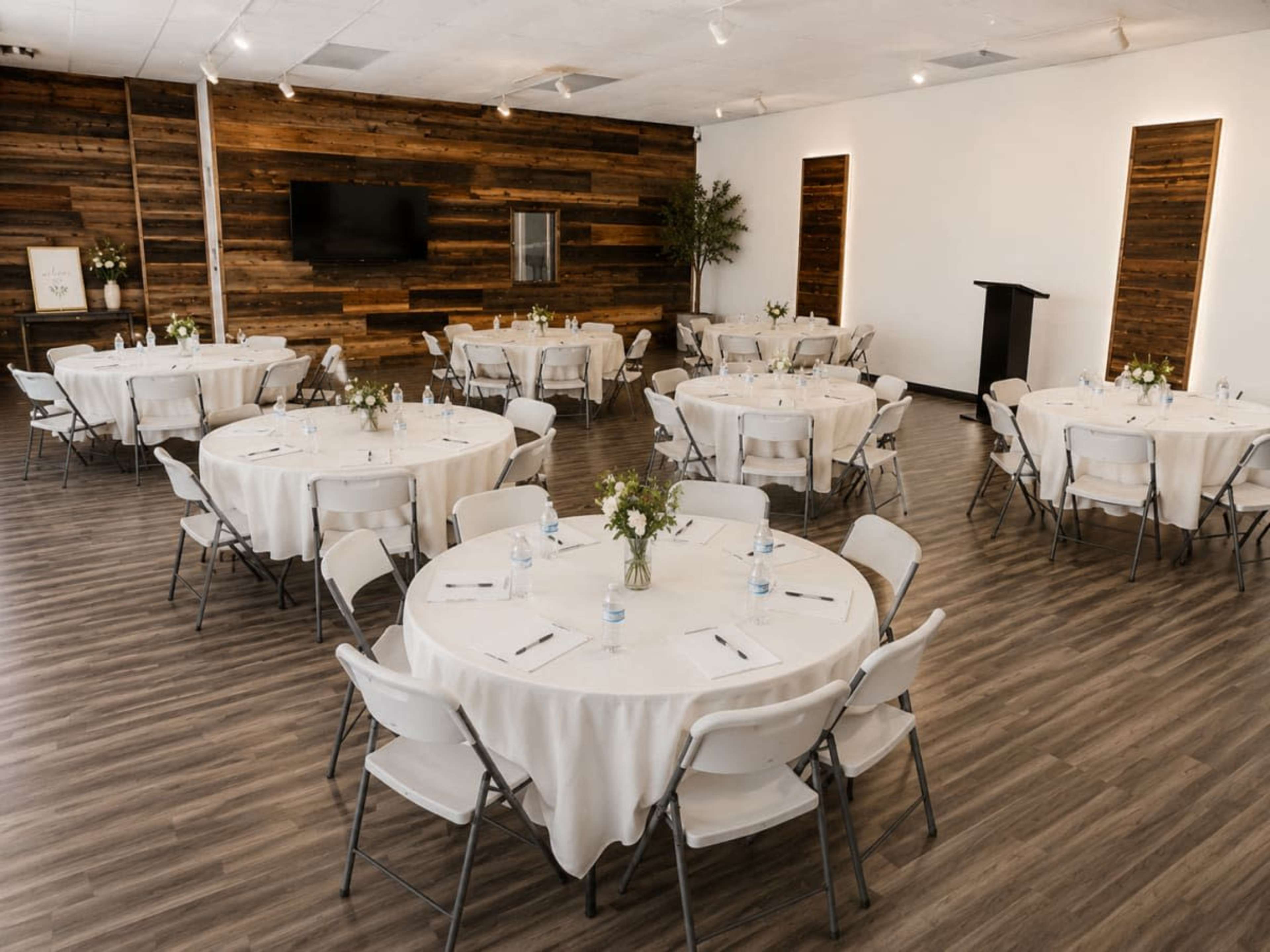 The image features a large indoor event space with multiple round tables covered in white tablecloths, each set with water bottles and utensils, surrounded by wooden walls and soft lighting.
