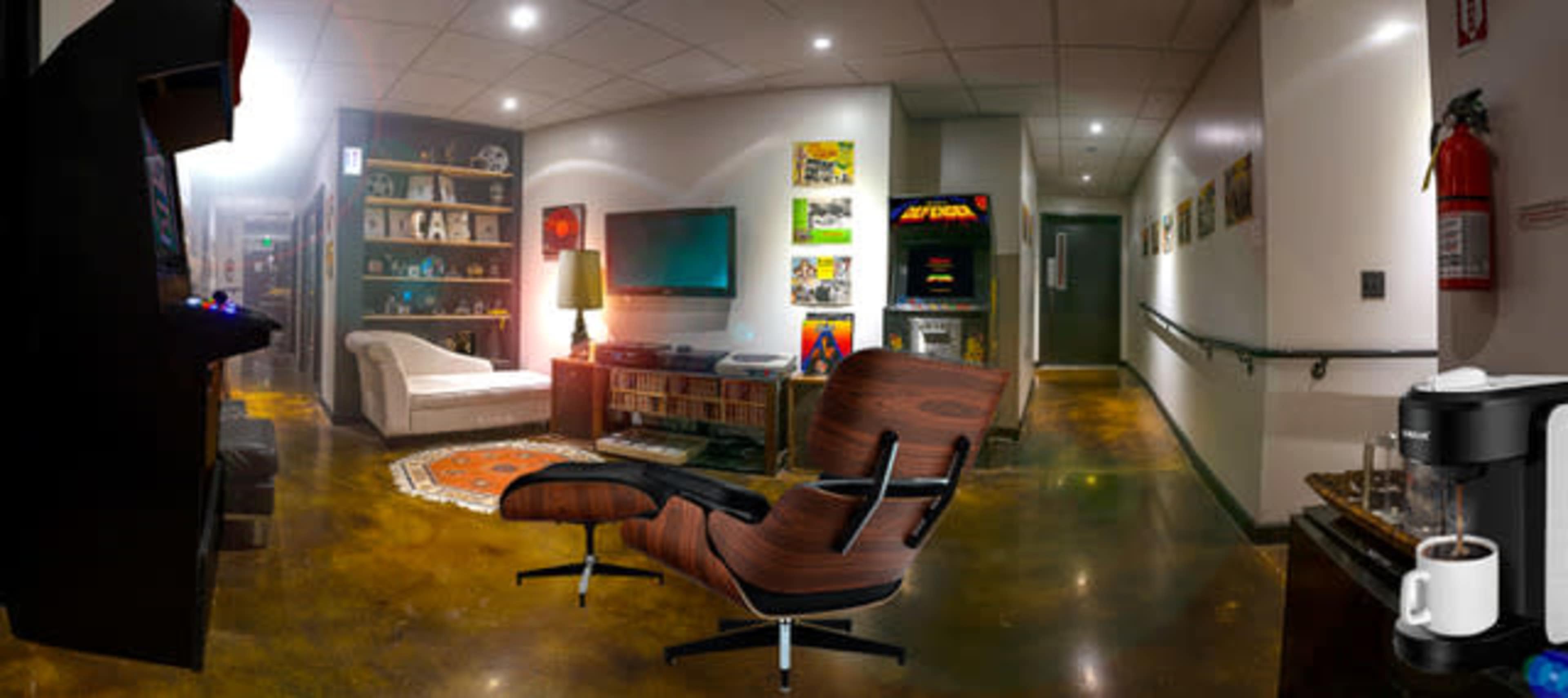 The image shows a modern lounge area with a comfortable chair, a gaming machine, a television, and a hallway with framed pictures and a coffee maker.