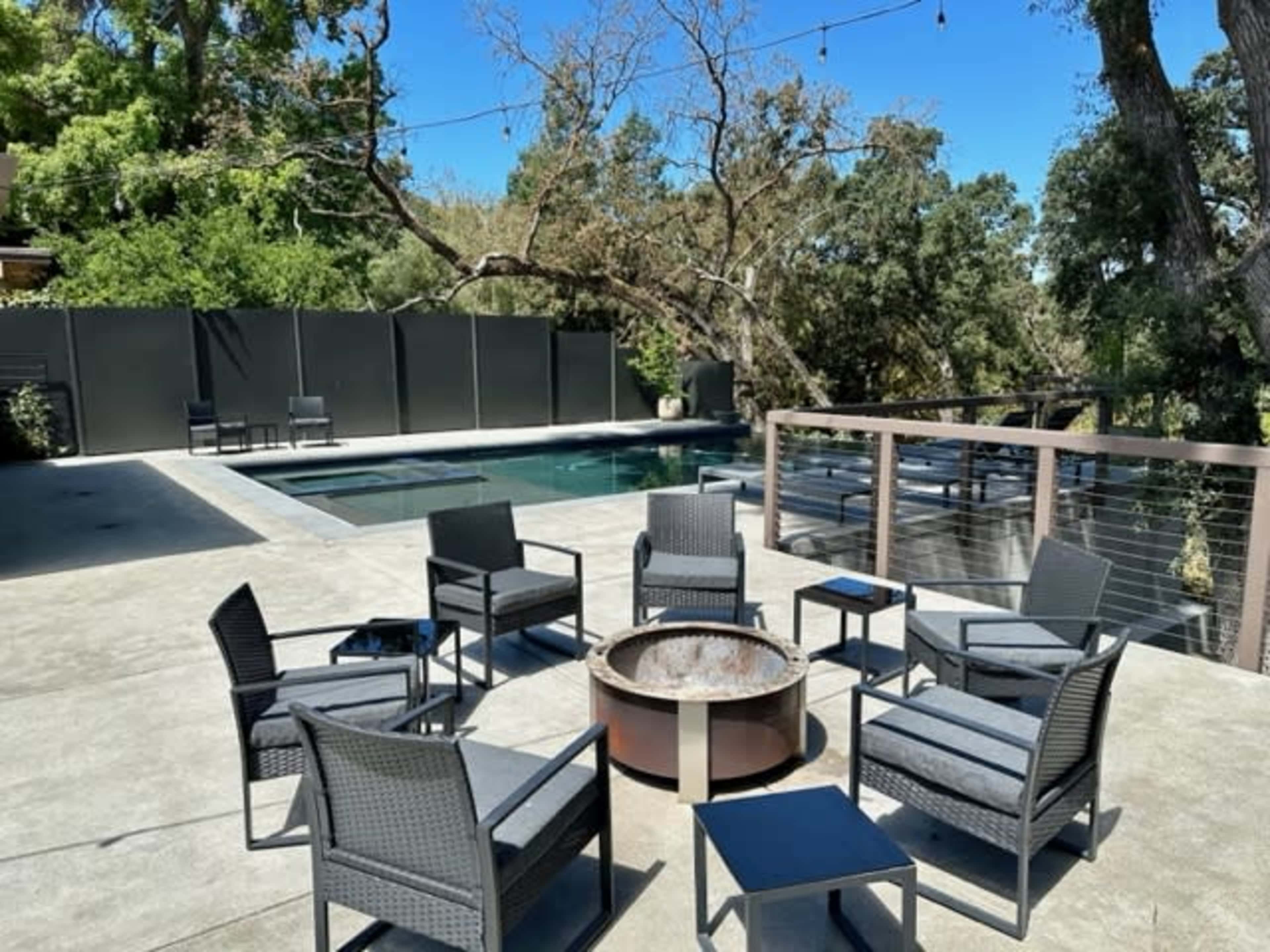 A seating area with six chairs arranged around a fire pit overlooks a pool surrounded by trees and a deck.