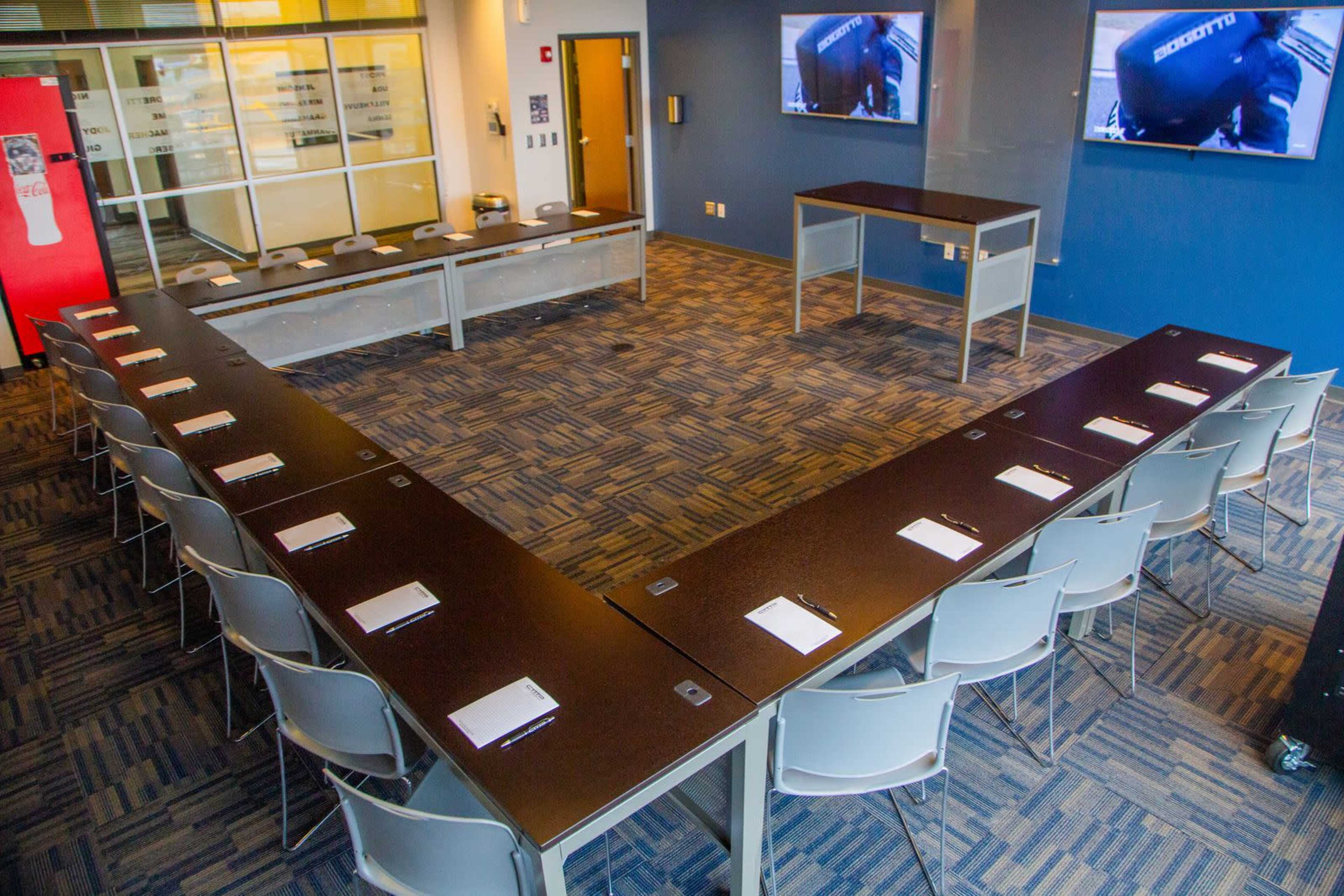 A conference room is set up in a U-shape with several tables and chairs, featuring monitors on the walls.
