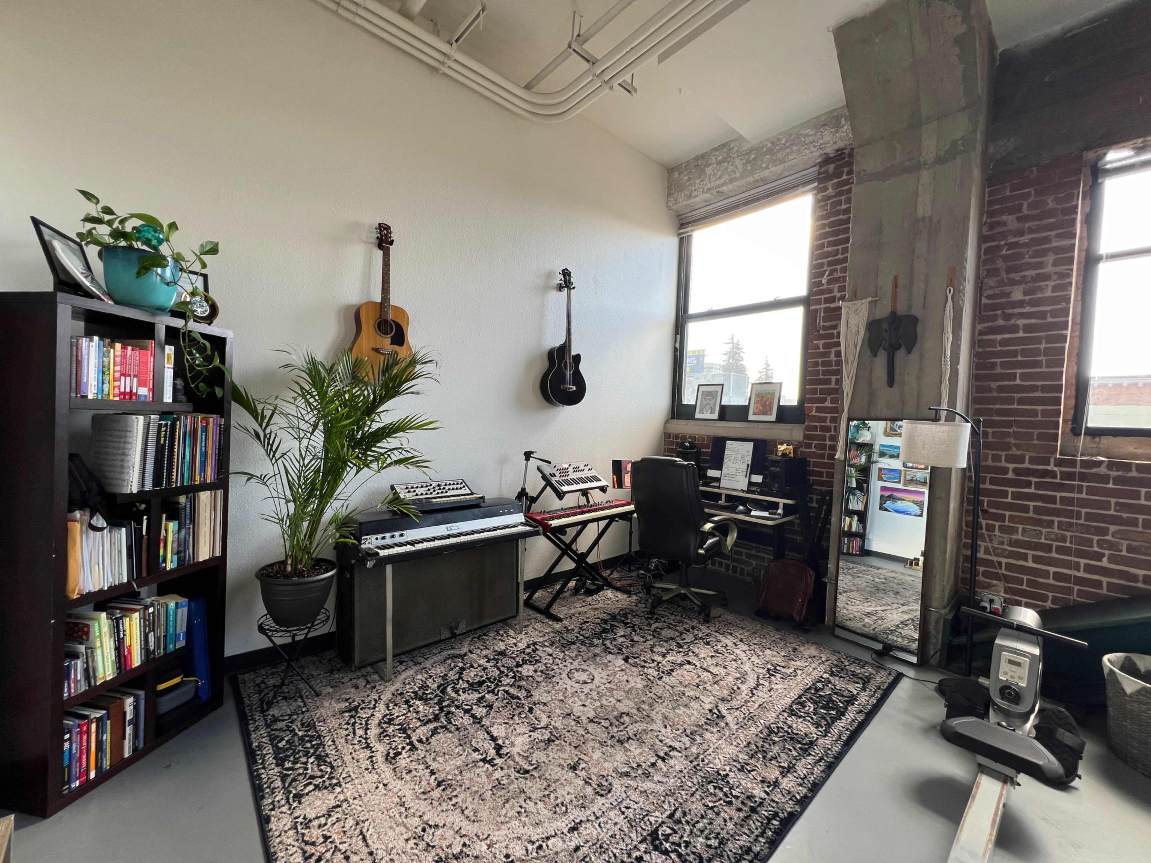 The image shows a workspace in a room featuring a keyboard, a guitar, a bookshelf filled with books, a plant, and a large mirror next to a window.