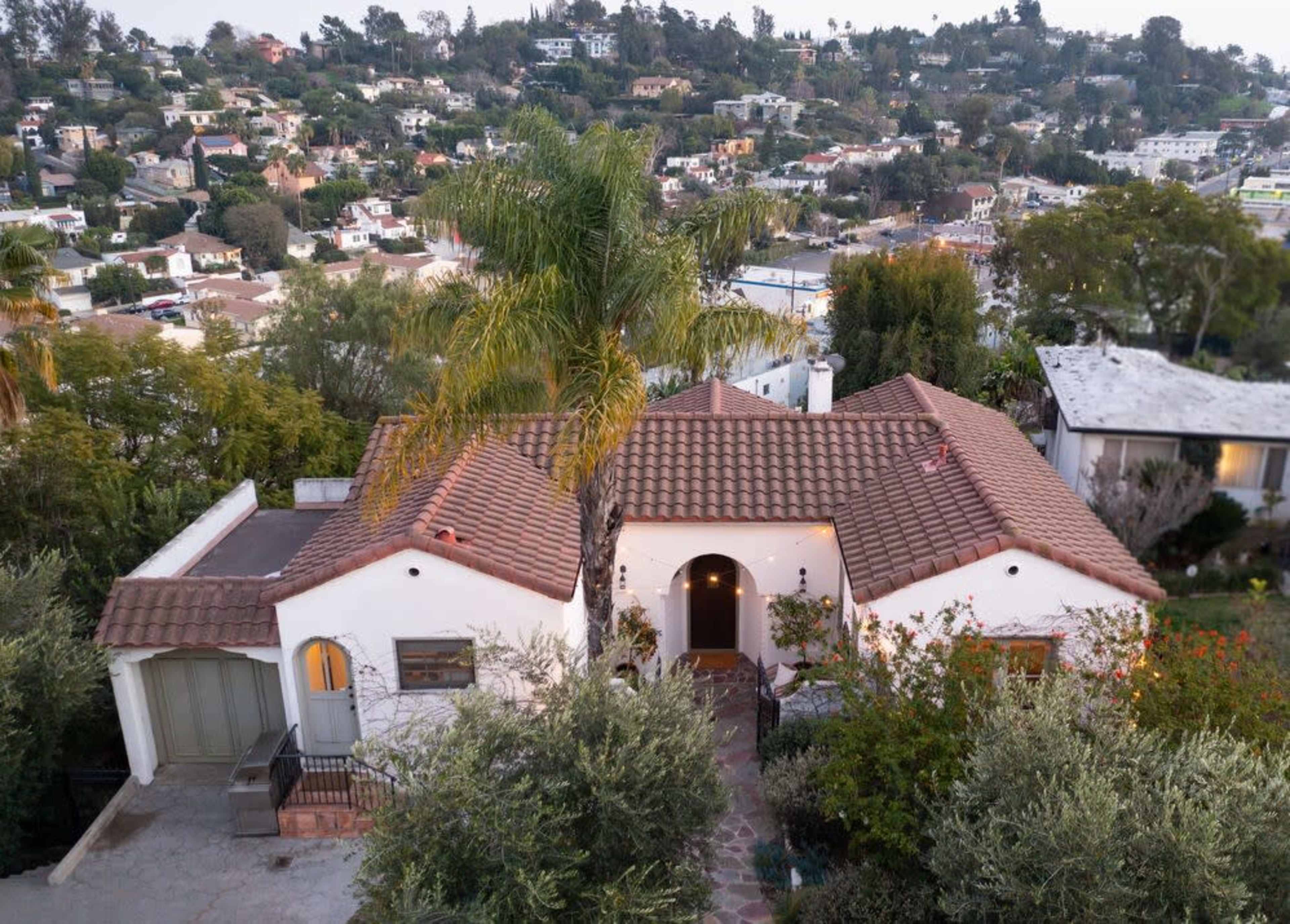 The image shows a single-story house with a tiled roof, surrounded by greenery and elevated on a hillside overlooking a neighborhood.