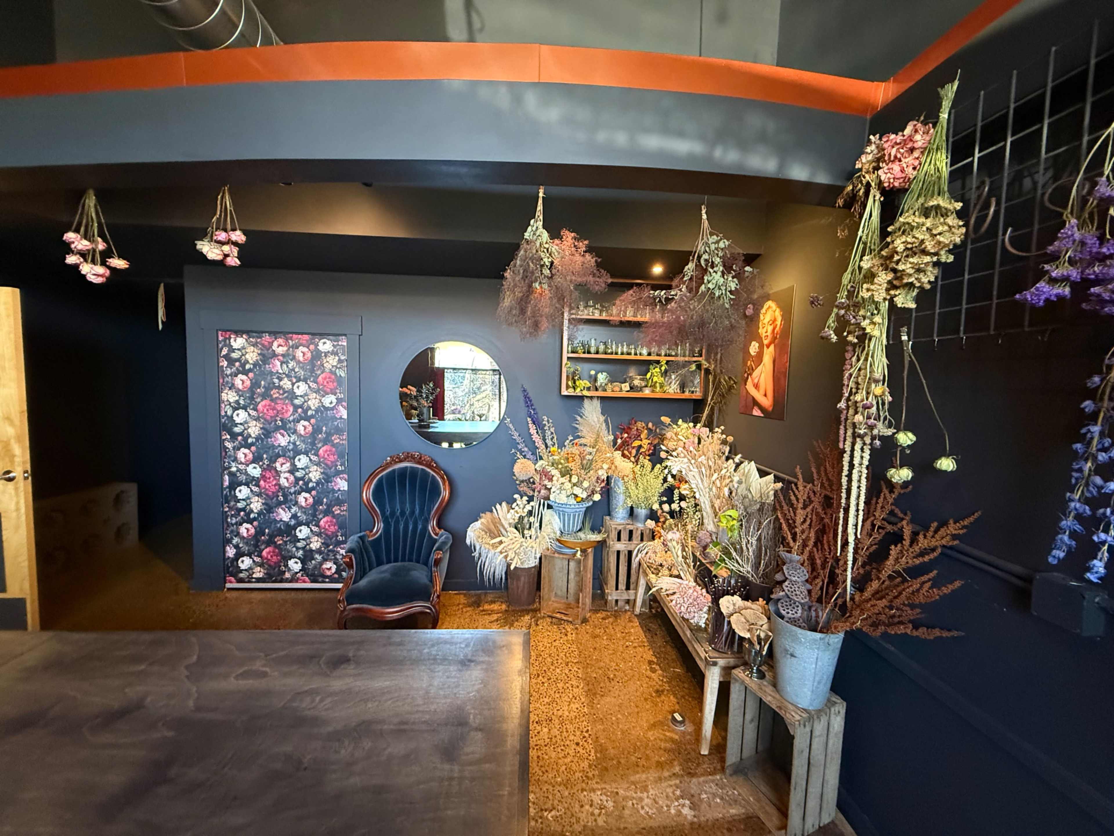 The image shows a cozy, decorated corner of a room featuring a vintage chair, a round mirror, and various dried flowers arranged in rustic wooden crates.
