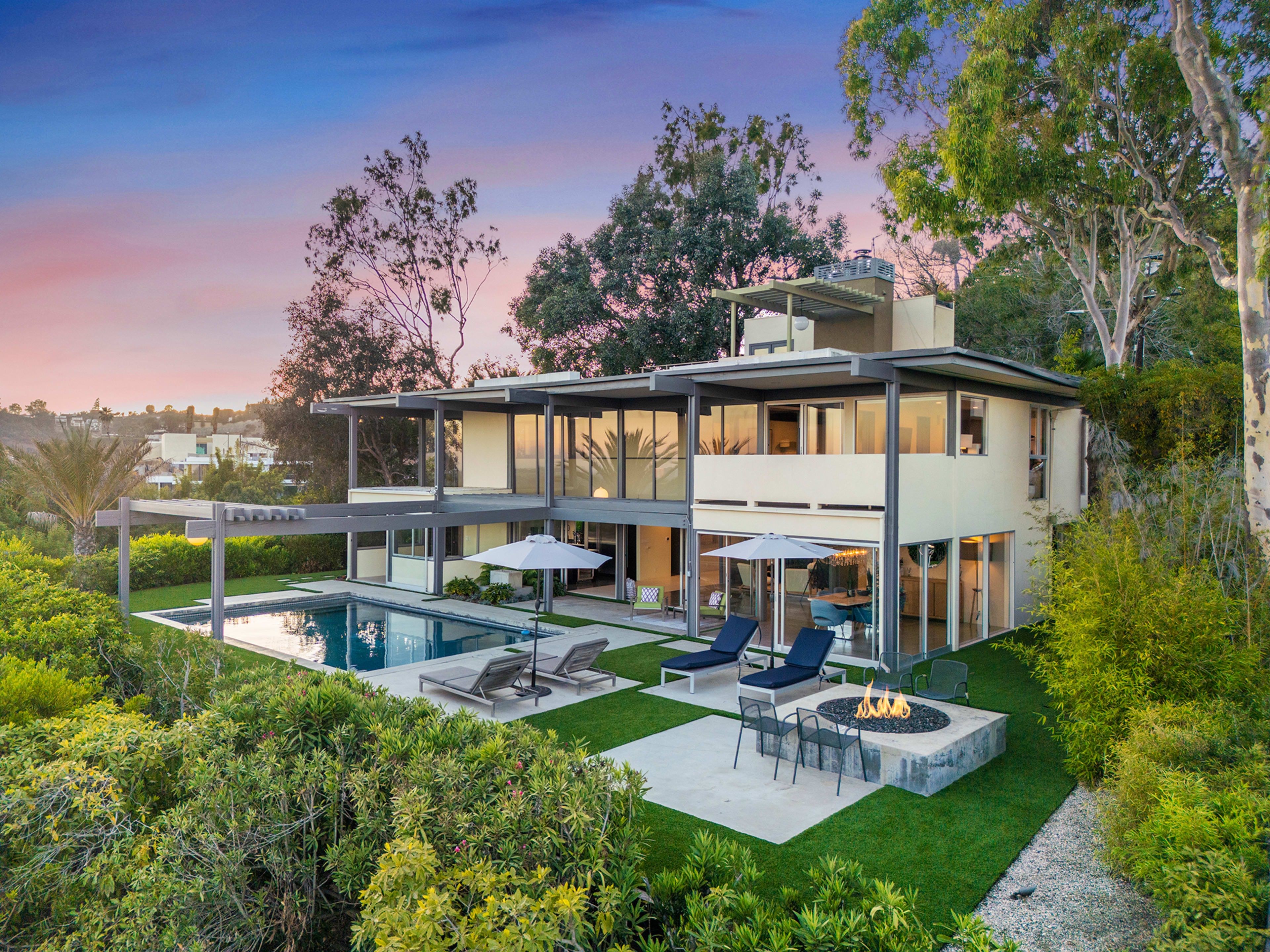 A modern two-story house with large glass windows overlooks a pool and fire pit, surrounded by greenery and trees.