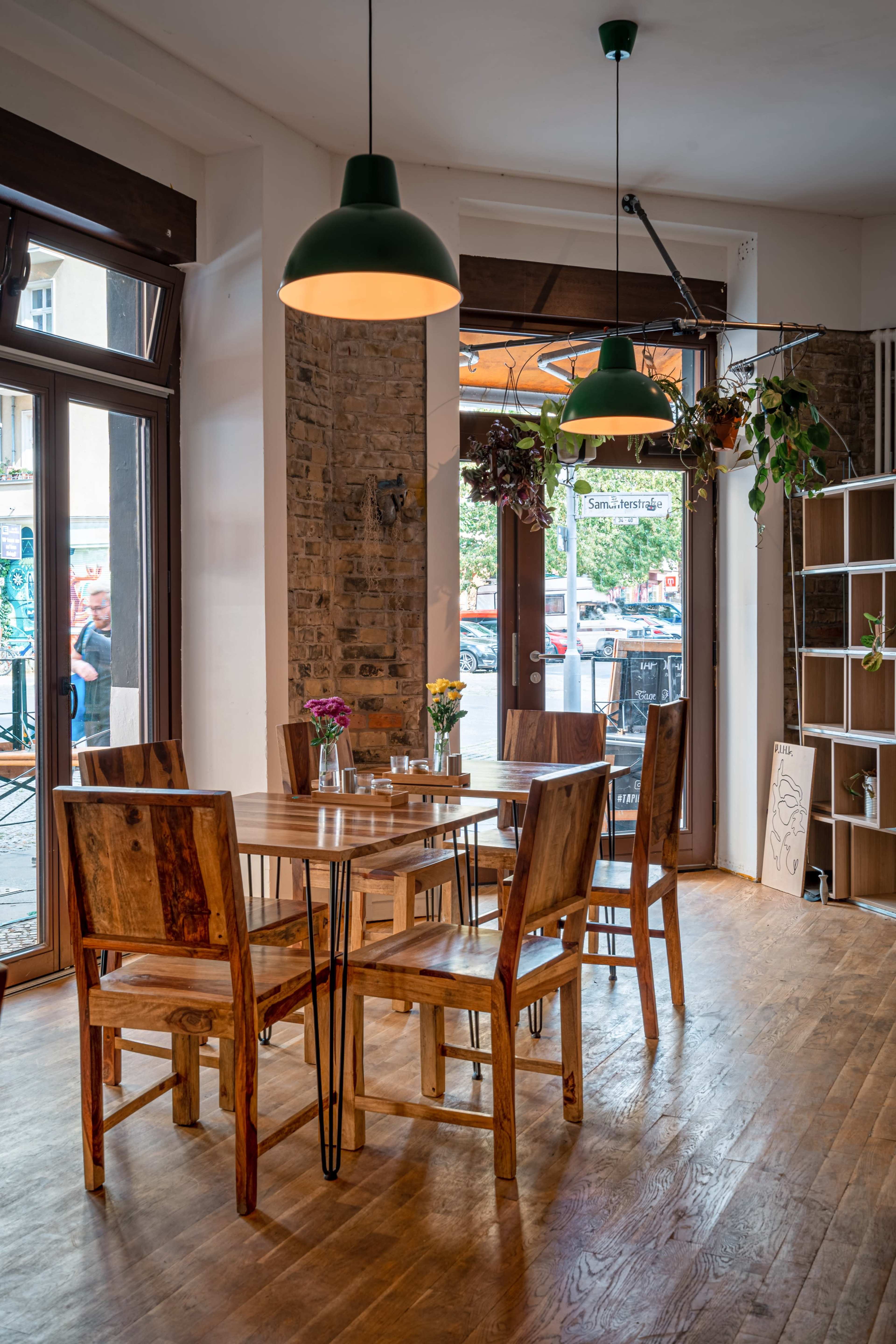 A wooden table surrounded by five chairs is placed near large windows with plants hanging above in a bright cafe.