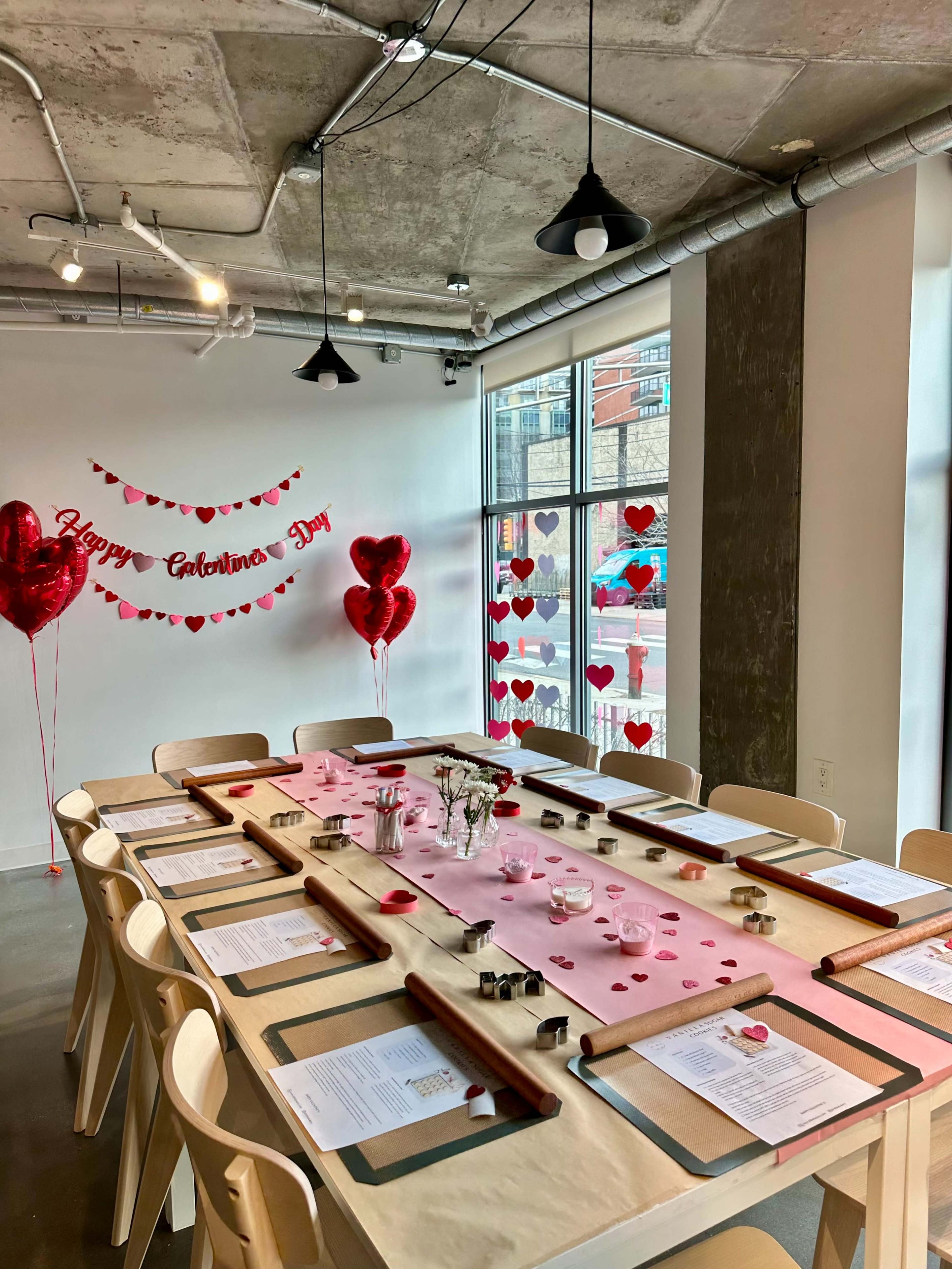 A festive dining setup is arranged with a long table covered in pink paper, surrounded by heart-shaped decorations and balloons for Valentine's Day.