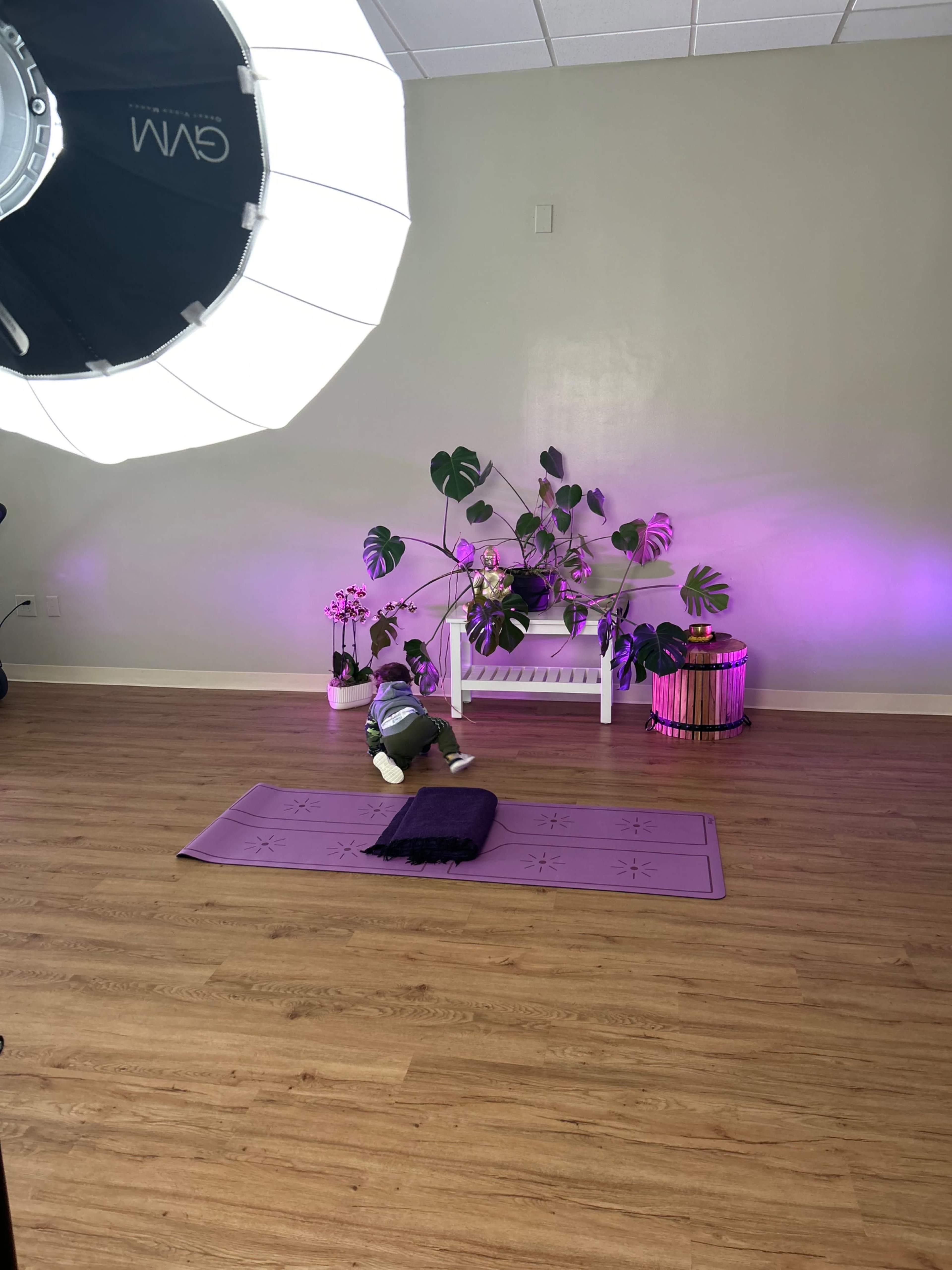 A child sits on a yoga mat in a studio with plants and soft purple lighting.