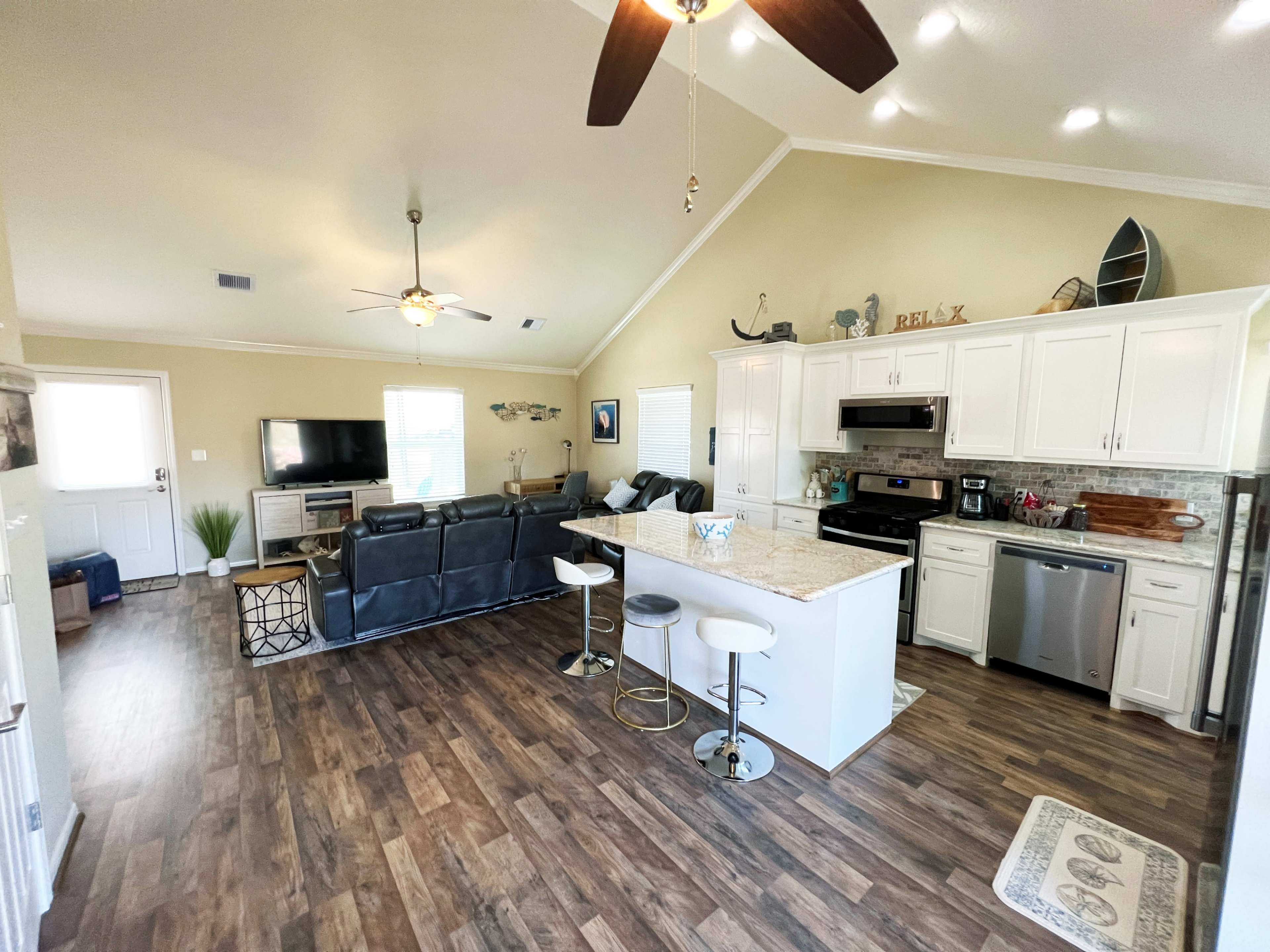 The image shows a modern open-concept living area with a kitchen, featuring white cabinets, a central island with seating, and a living room with a large television and seating arrangement.