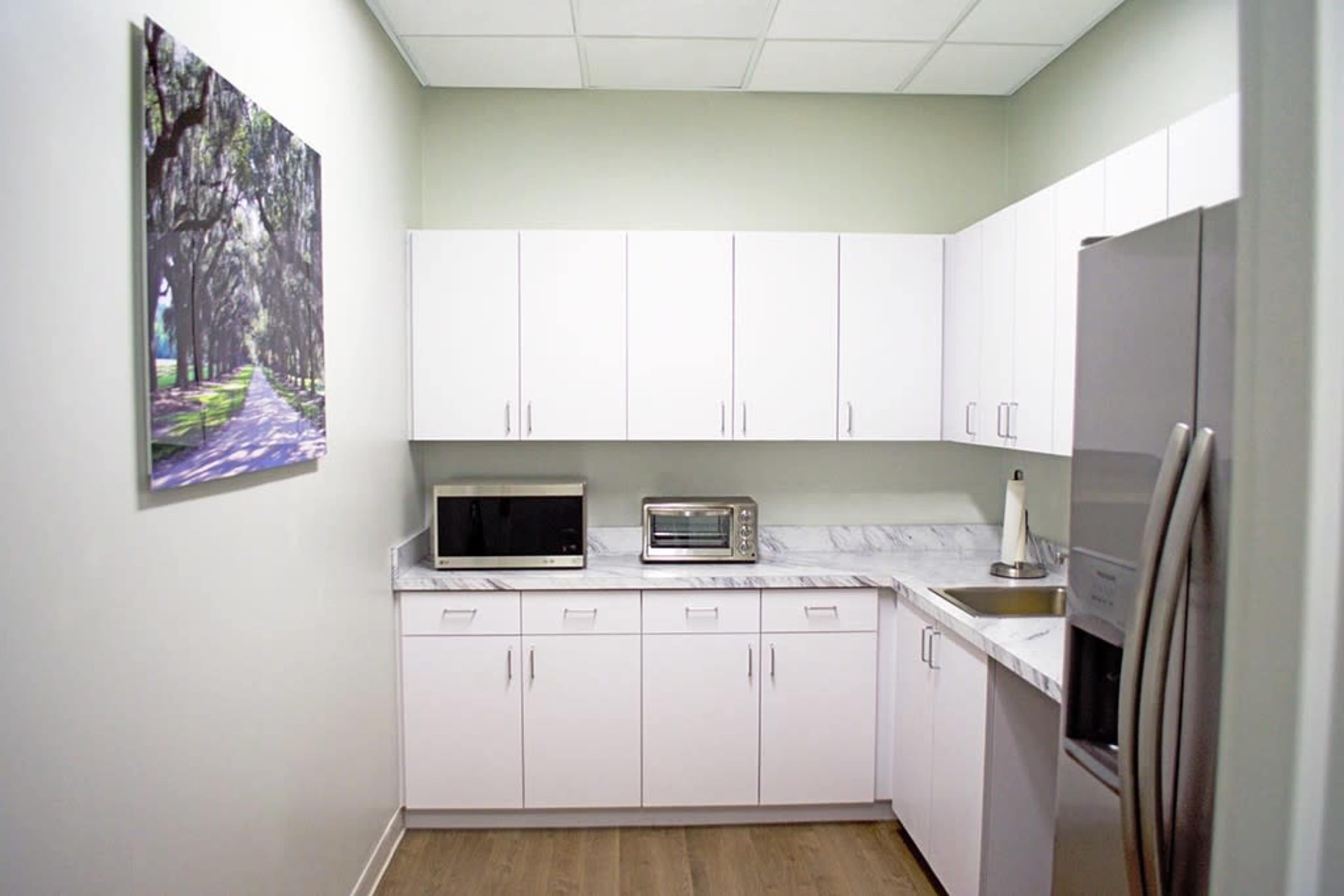 The image shows a small kitchen area with white cabinets, a marble countertop, a microwave, a toaster oven, and a stainless steel refrigerator.