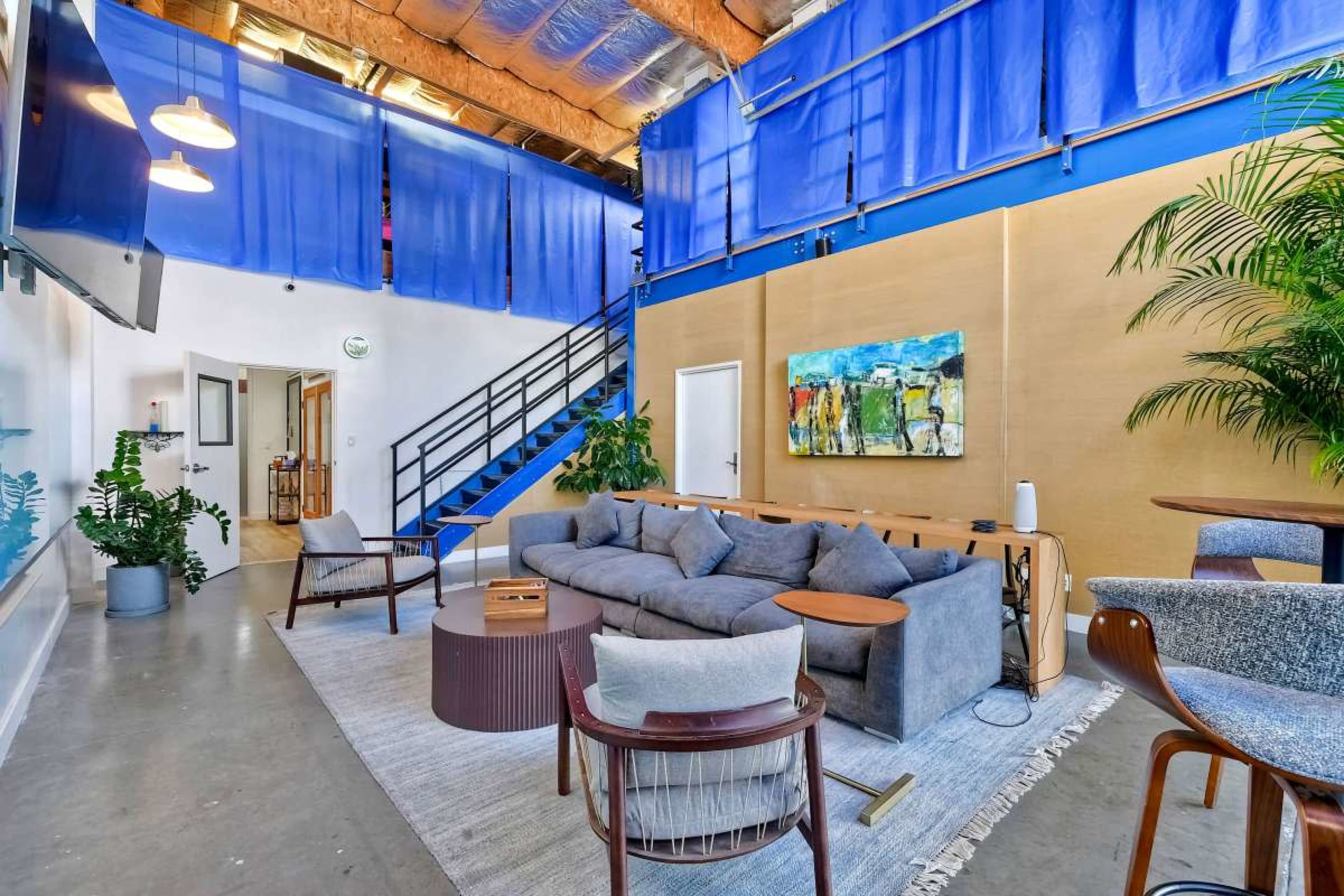 The image shows a modern living area featuring a large gray sectional sofa, a low coffee table, and a staircase leading to an upper level, with blue curtains draping above.