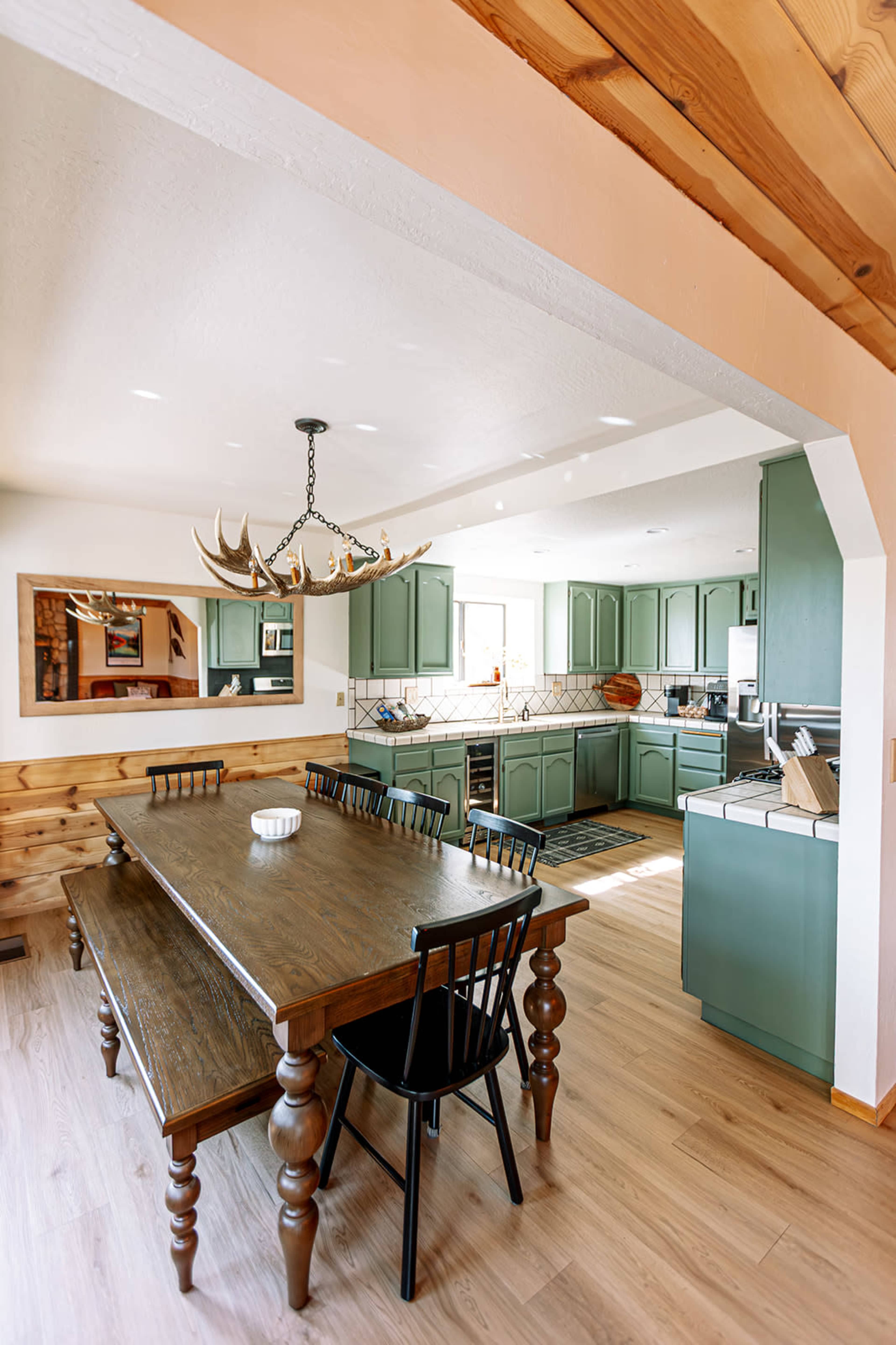 The image shows a kitchen and dining area featuring a wooden table with black chairs, green cabinetry, and a chandelier with antlers.