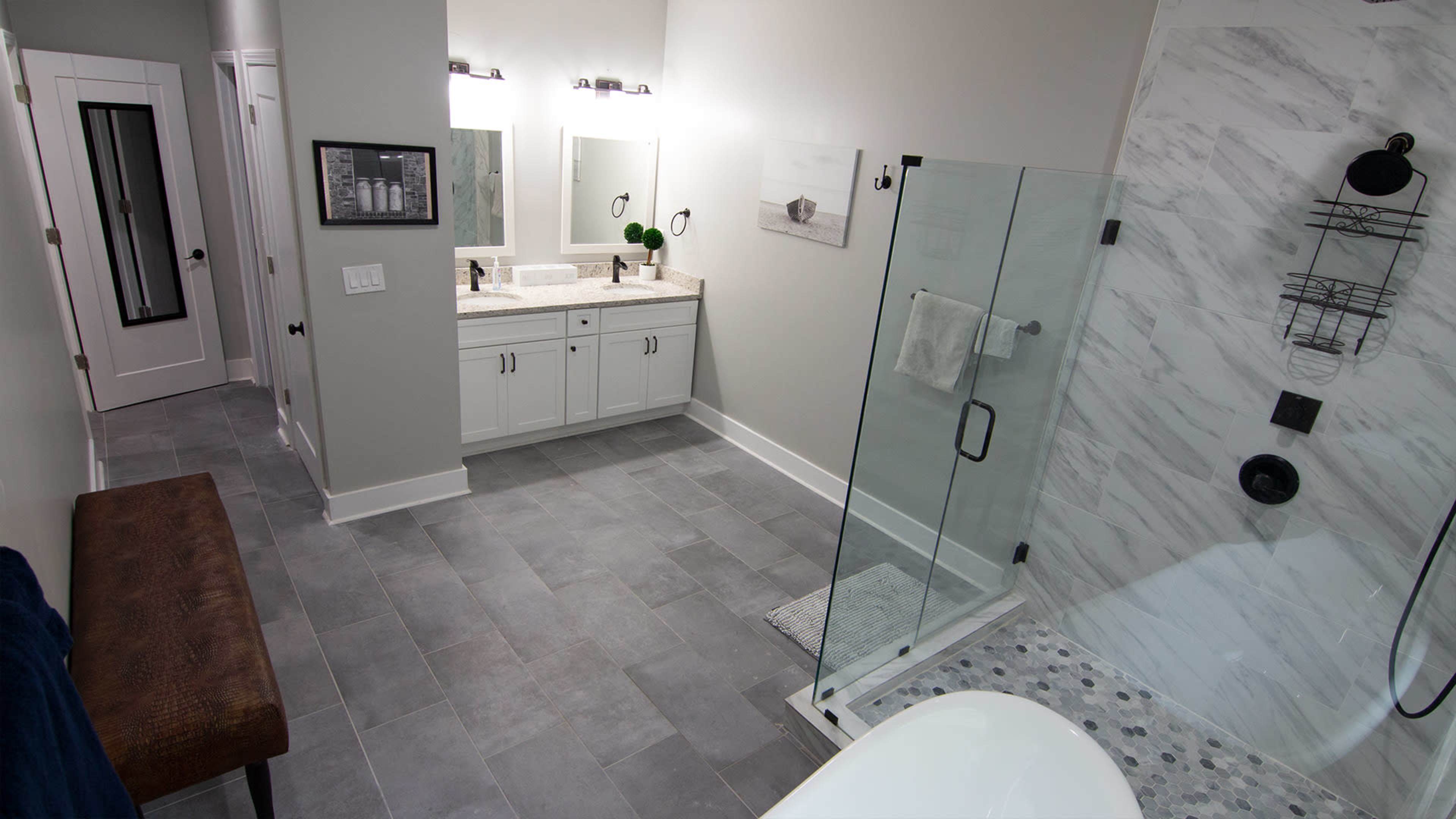 A modern bathroom with a double vanity, glass shower enclosure, and freestanding bathtub, featuring light-colored walls and gray tile flooring.