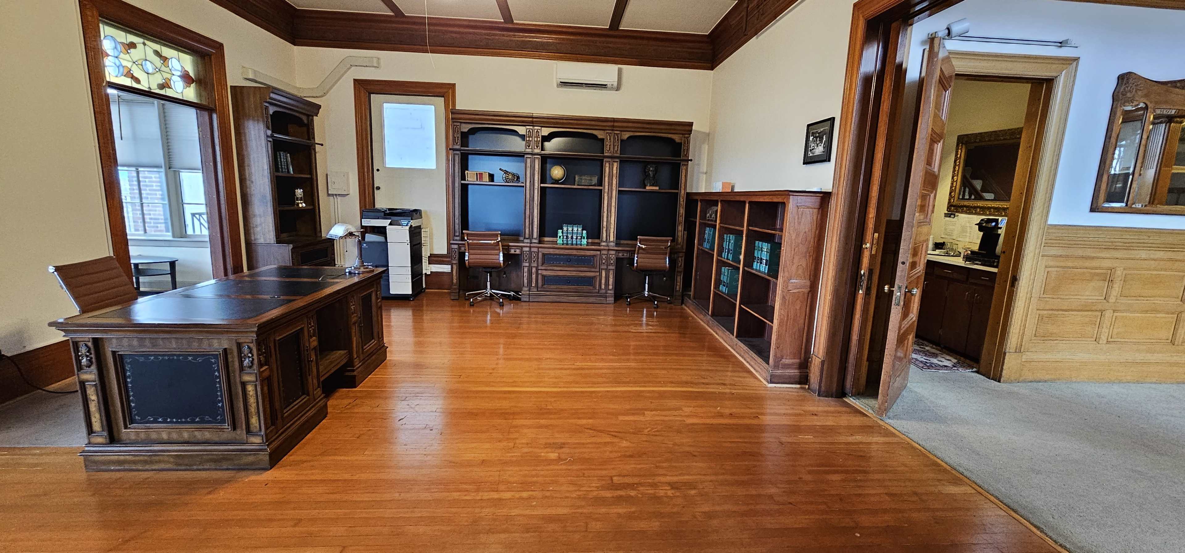 A spacious office with wooden flooring, a large dark wooden desk, shelves filled with books, and a doorway leading to another room.