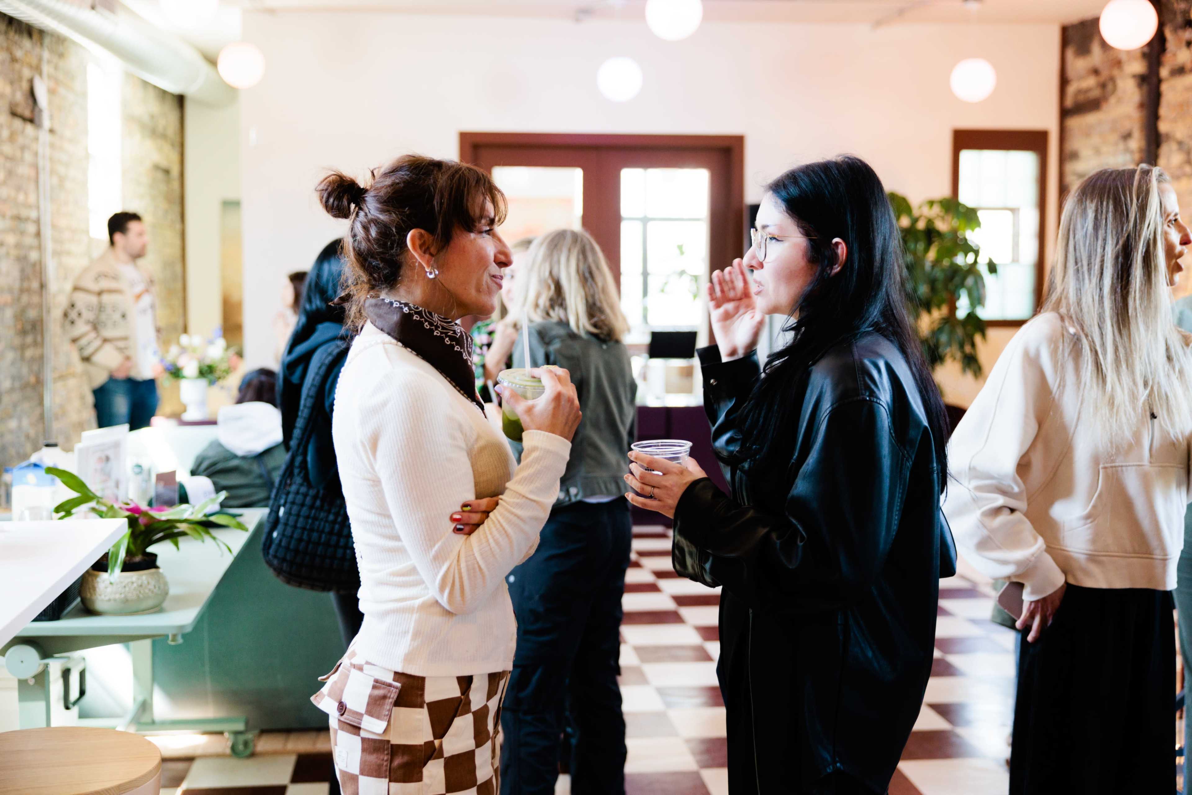 Two women engage in conversation while holding drinks in a brightly lit room with people socializing in the background.