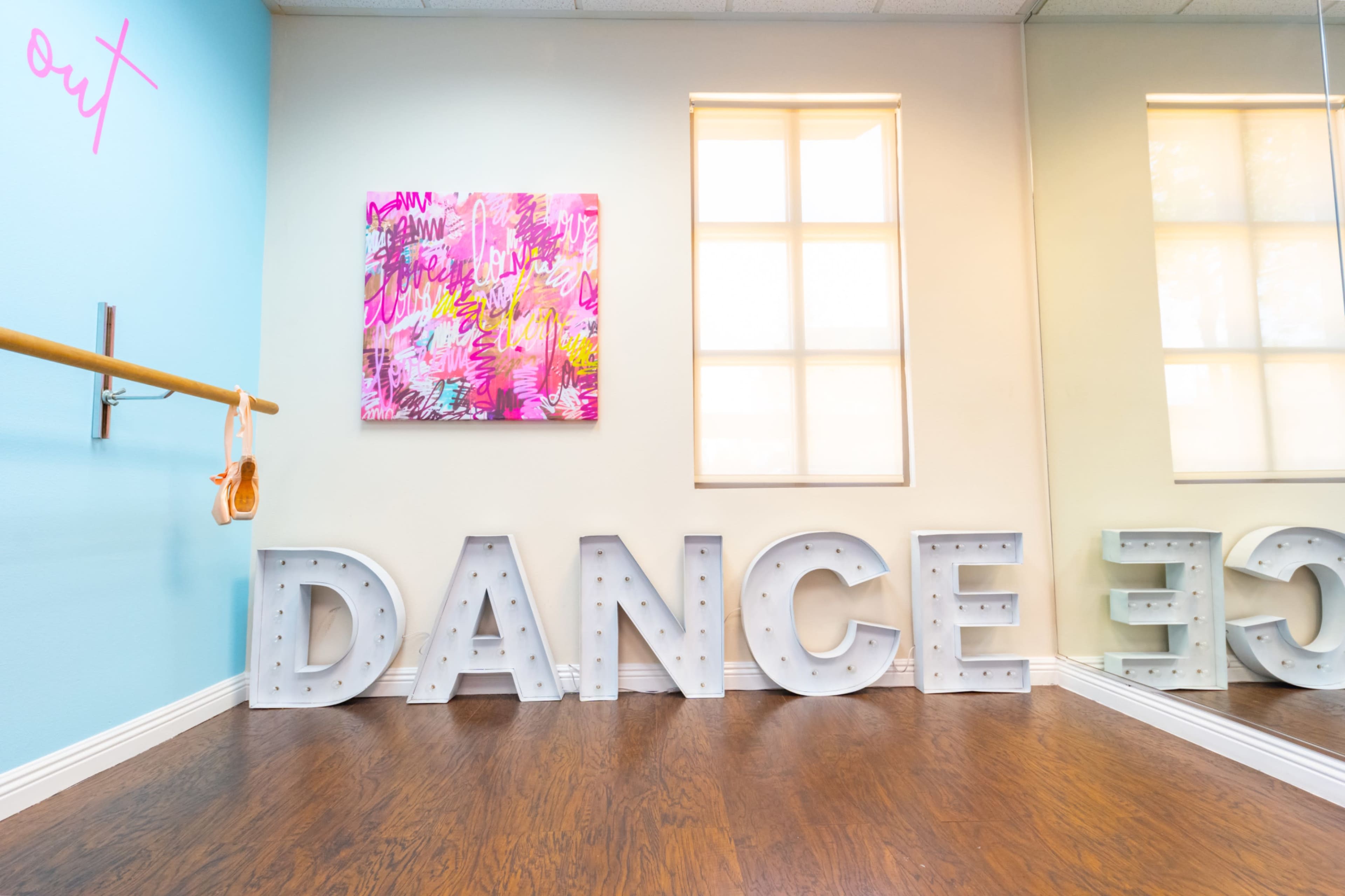 The image shows a dance studio with a wooden floor, colorful artwork on the wall, and illuminated letters spelling "DANCE."