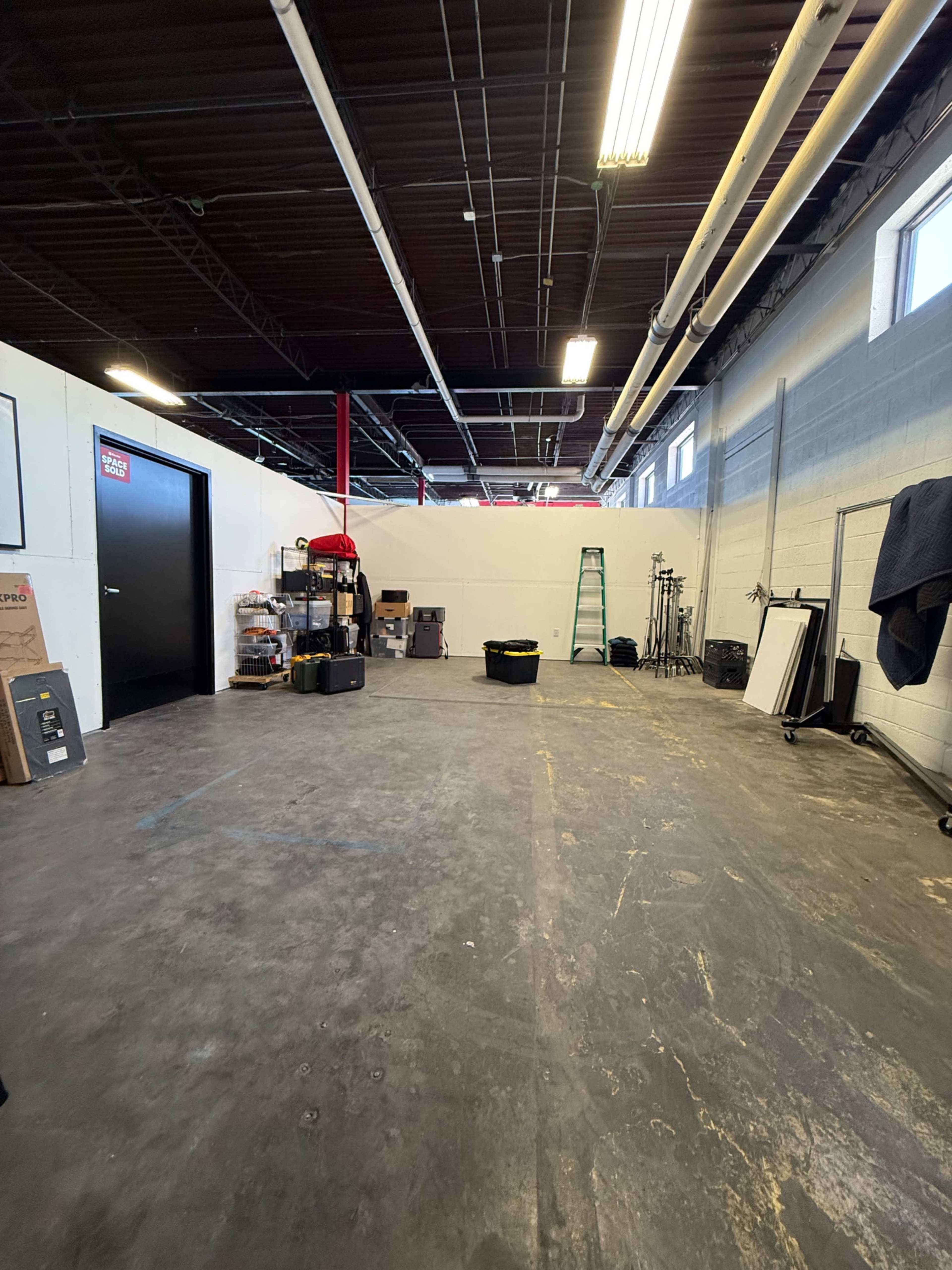 The image shows a spacious industrial-style room with a concrete floor, various storage items, and equipment along the walls, including a ladder and lighting setup.