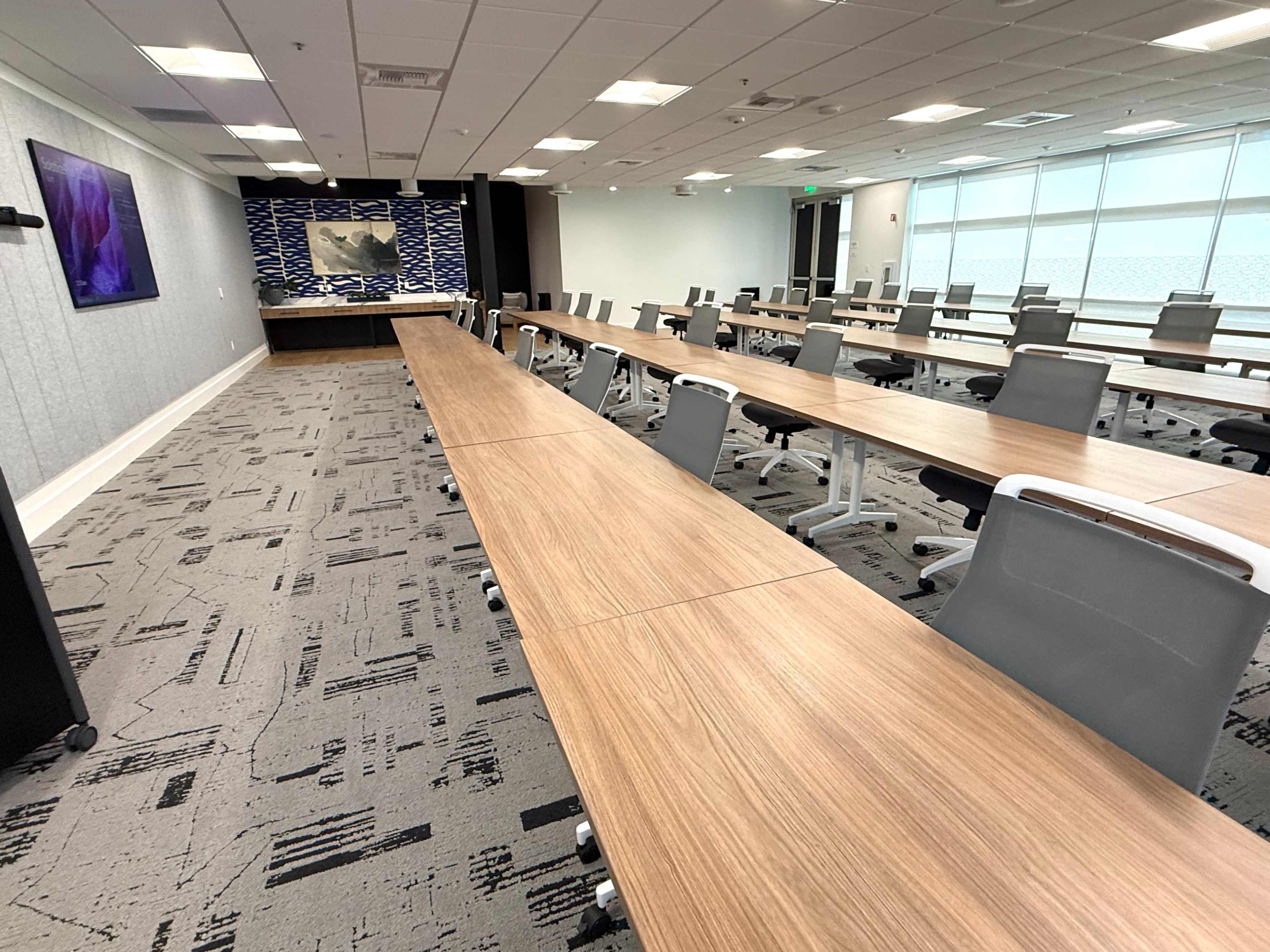 The image shows a modern conference room with long wooden tables arranged in a U-shape, equipped with ergonomic chairs and a presentation area at the front.