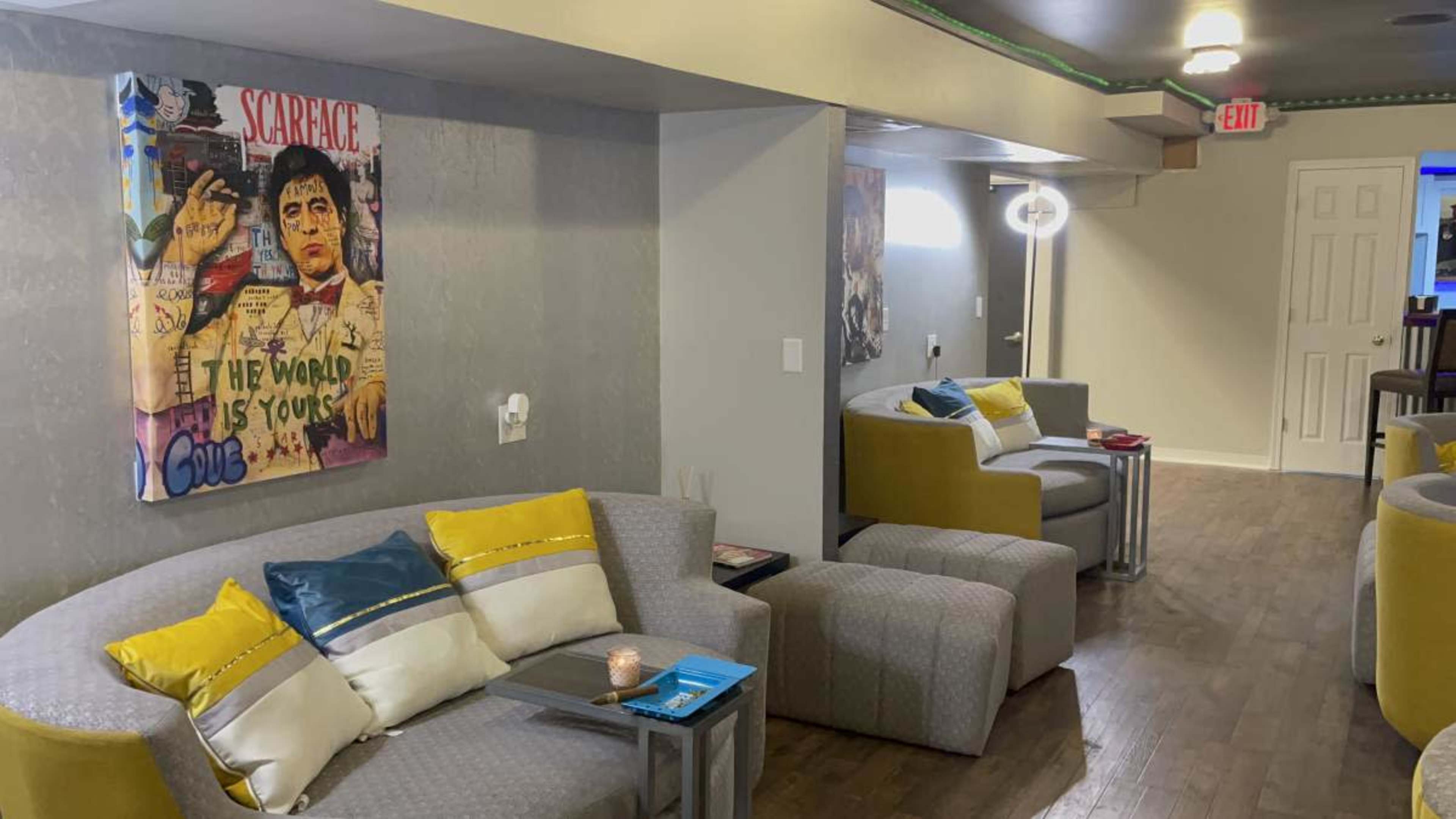 The image shows a contemporary lounge area with gray sofas adorned with colorful pillows, a wall featuring a framed "Scarface" poster, and softly lit corners.