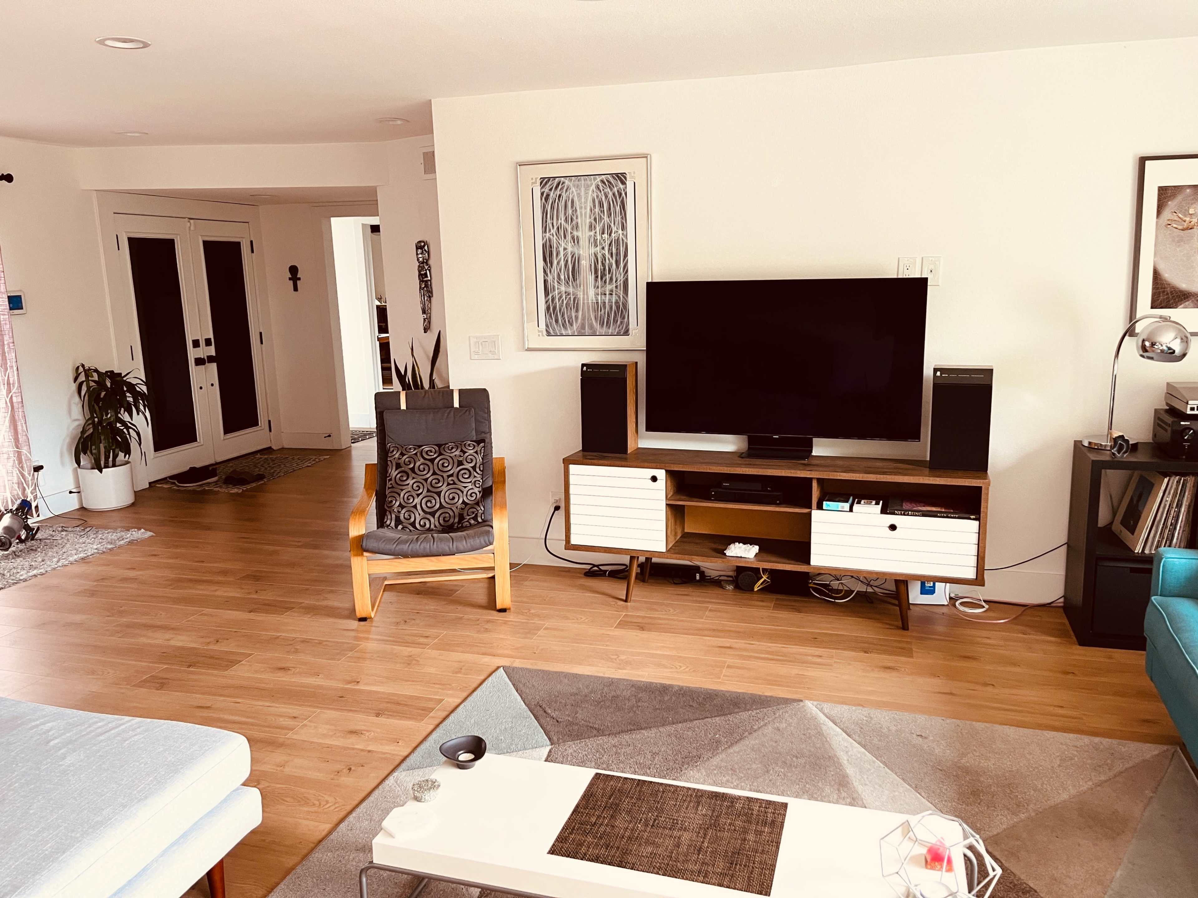 The living room features a television on a wooden cabinet with two speakers, surrounded by a blue couch and a patterned rug, with a visible entryway and potted plants in the background.