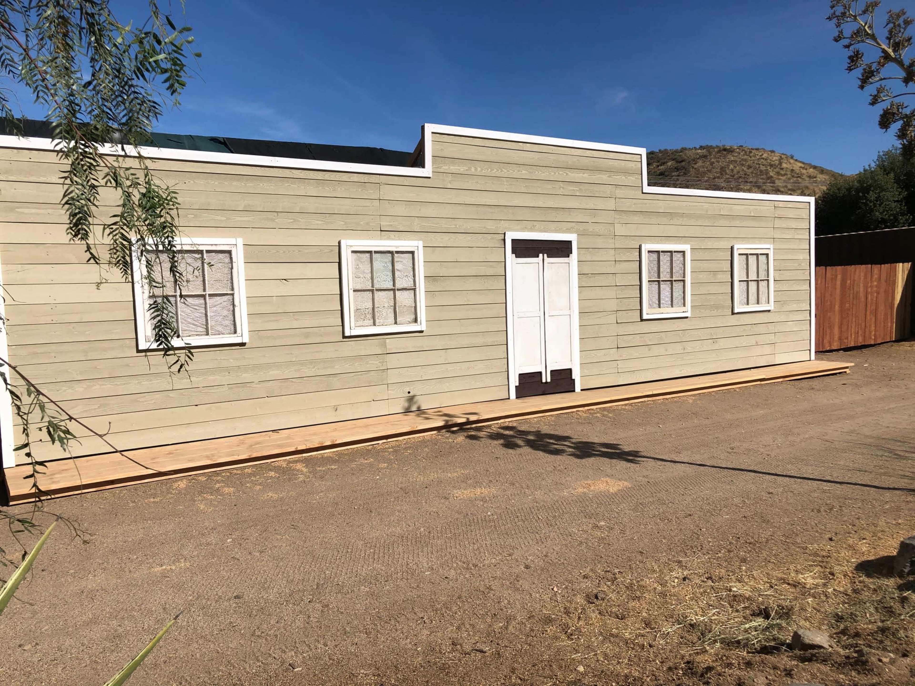 A rectangular wooden building with a light-colored exterior, accompanied by multiple windows and a central door, set against a clear blue sky and hillside.