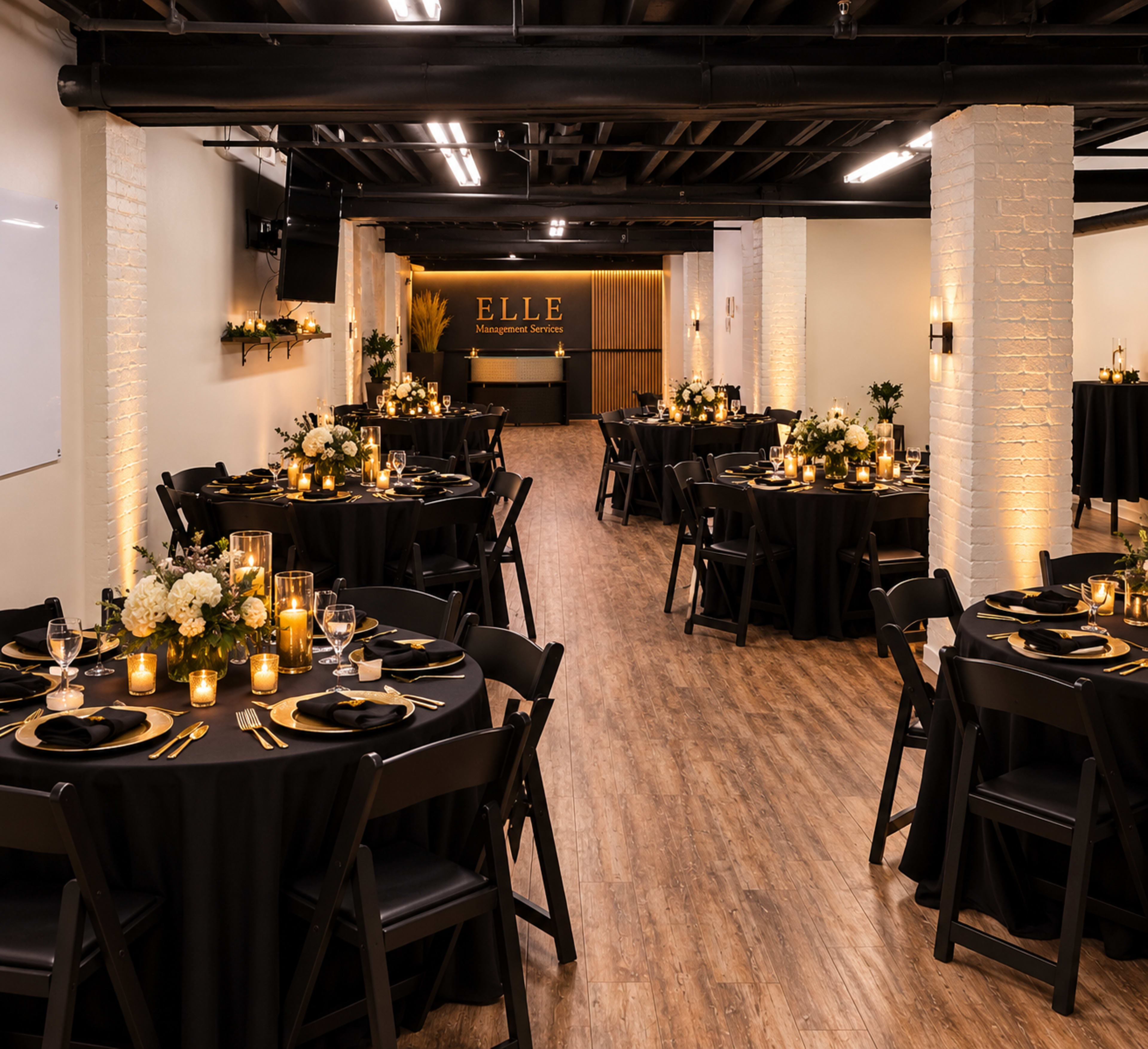 The image shows a stylish event space set up for dining, featuring round black tables adorned with floral centerpieces and candles, surrounded by a warm, inviting ambiance.