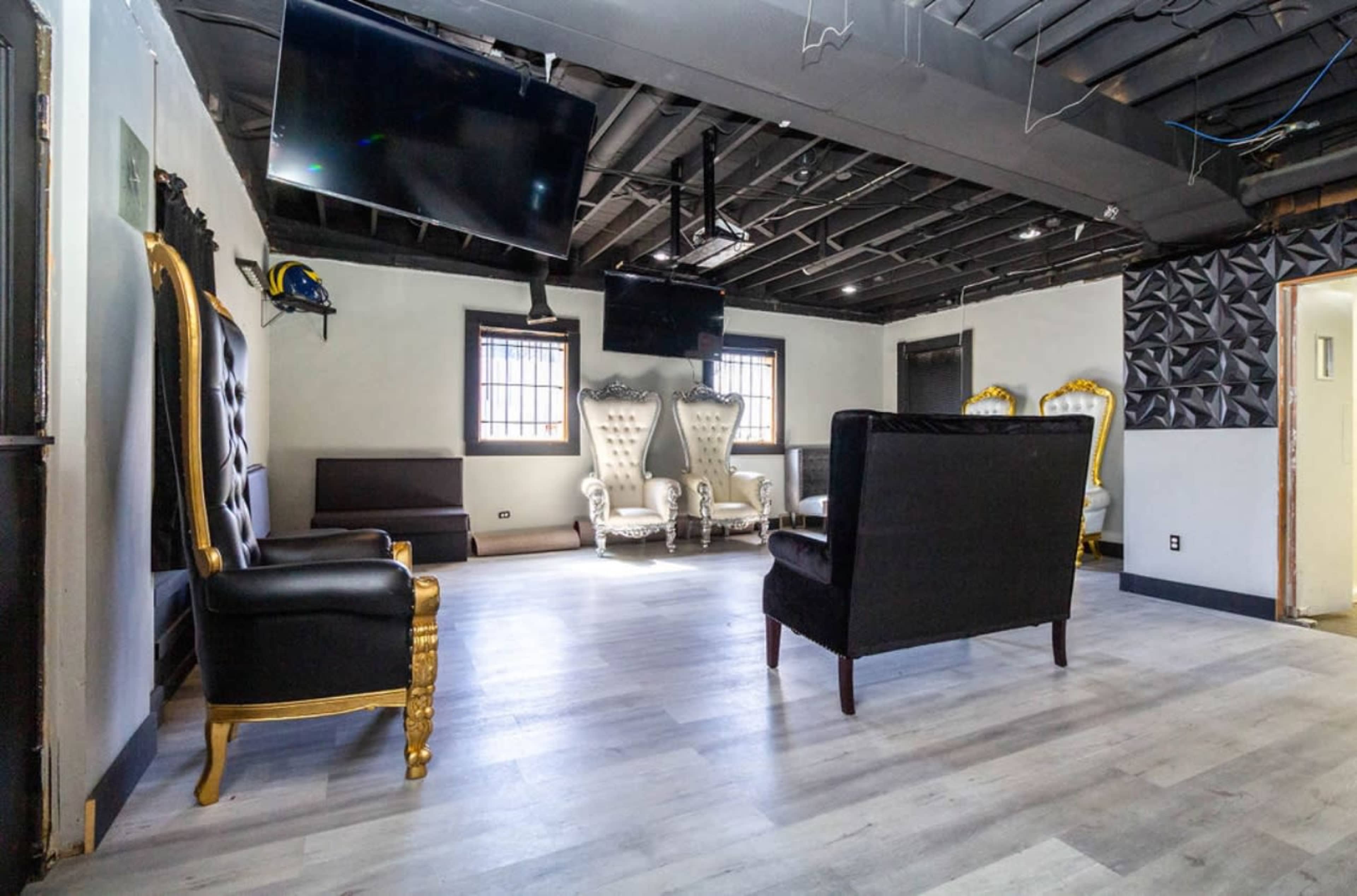 The room features several ornate chairs, two large flat-screen TVs on the walls, and modern flooring in a well-lit space with exposed ceiling beams.