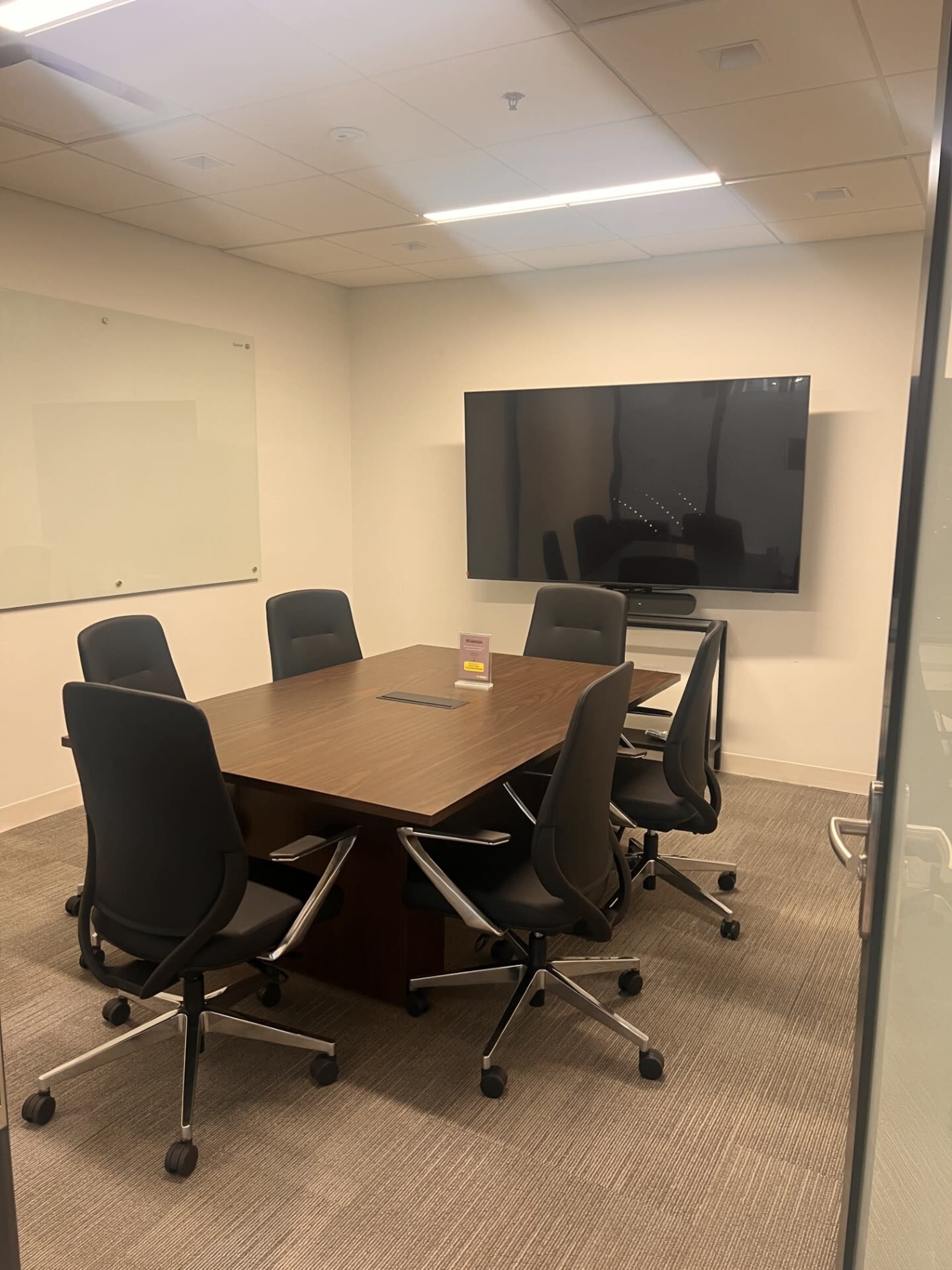 A modern conference room features a large wooden table surrounded by six black ergonomic chairs and a wall-mounted screen.