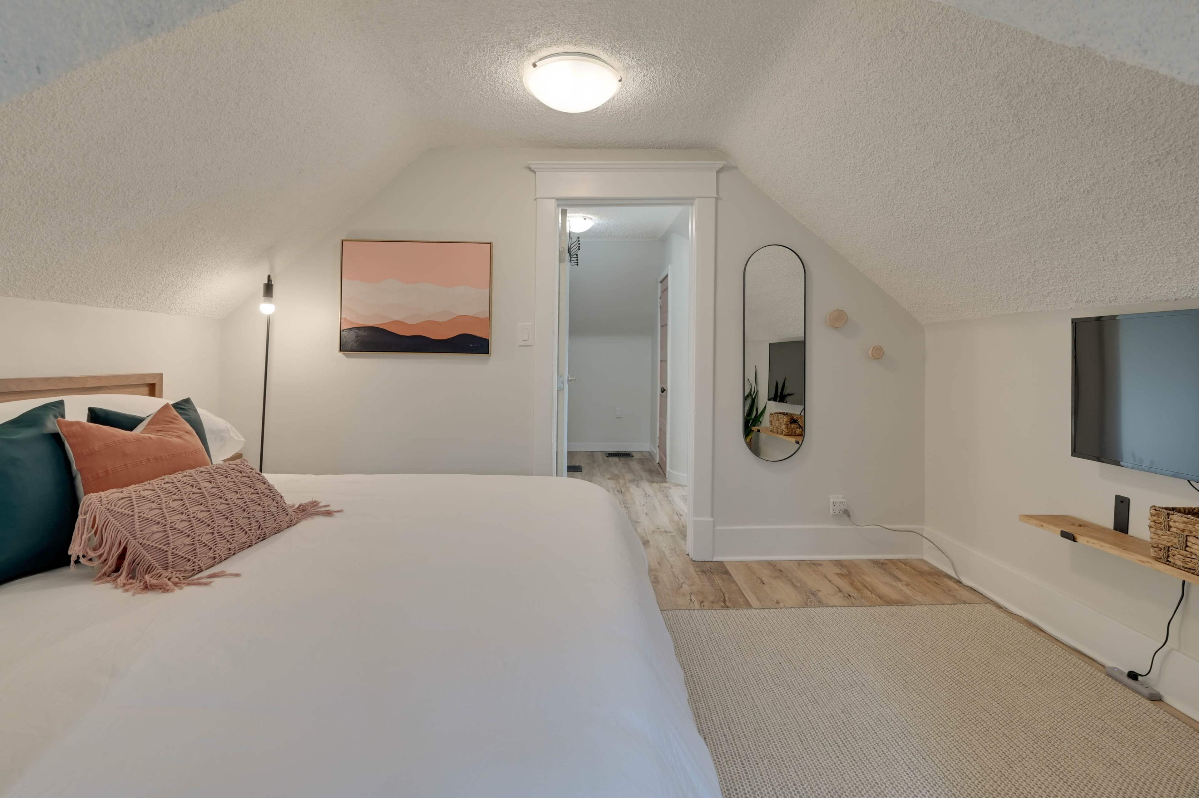 A cozy attic bedroom with a bed, decorative pillows, a wall-mounted mirror, and a small TV mounted on the wall.