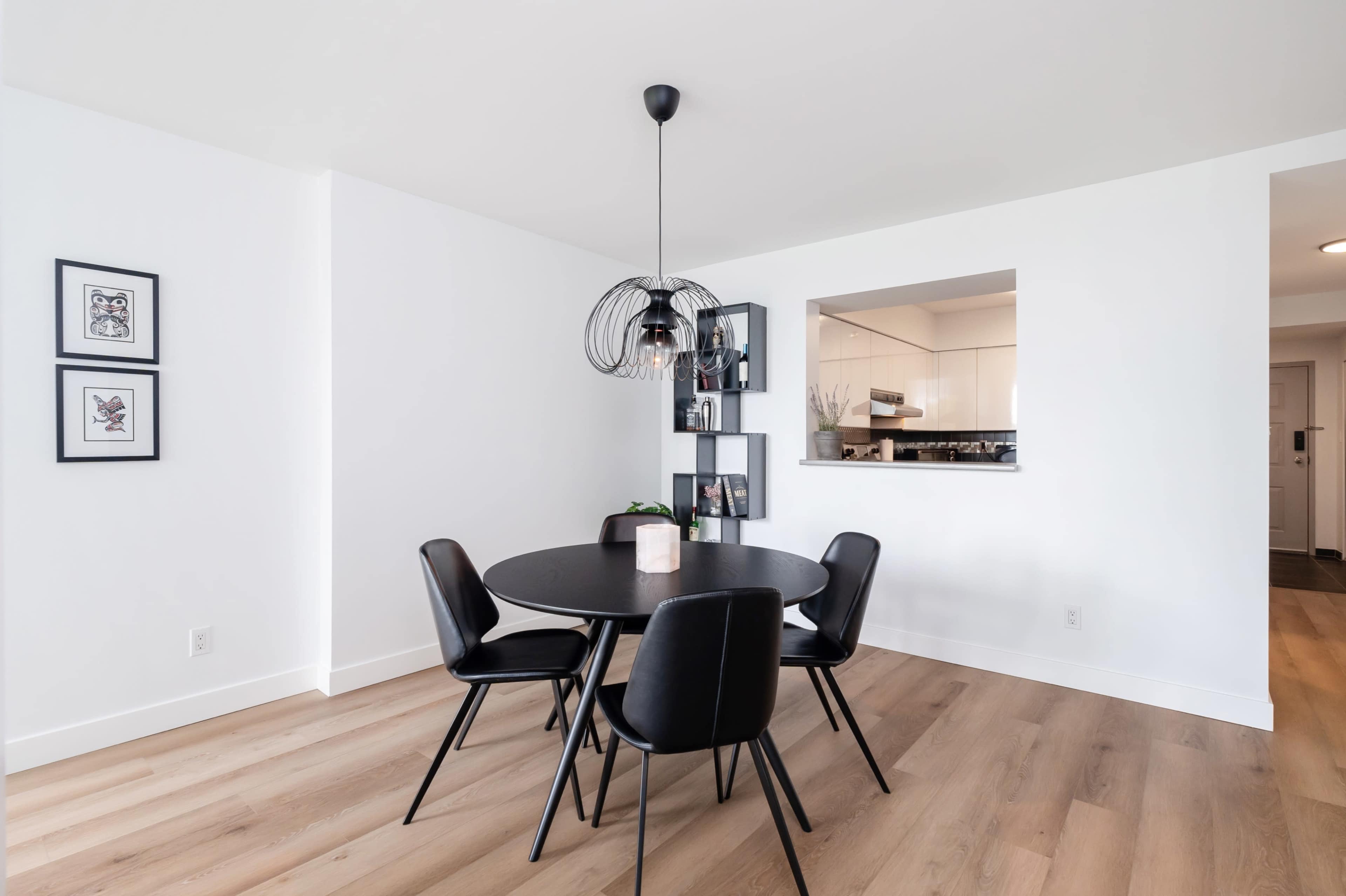 A modern dining area features a round black table surrounded by four black chairs, with a pendant light overhead and a view into an adjacent kitchen.