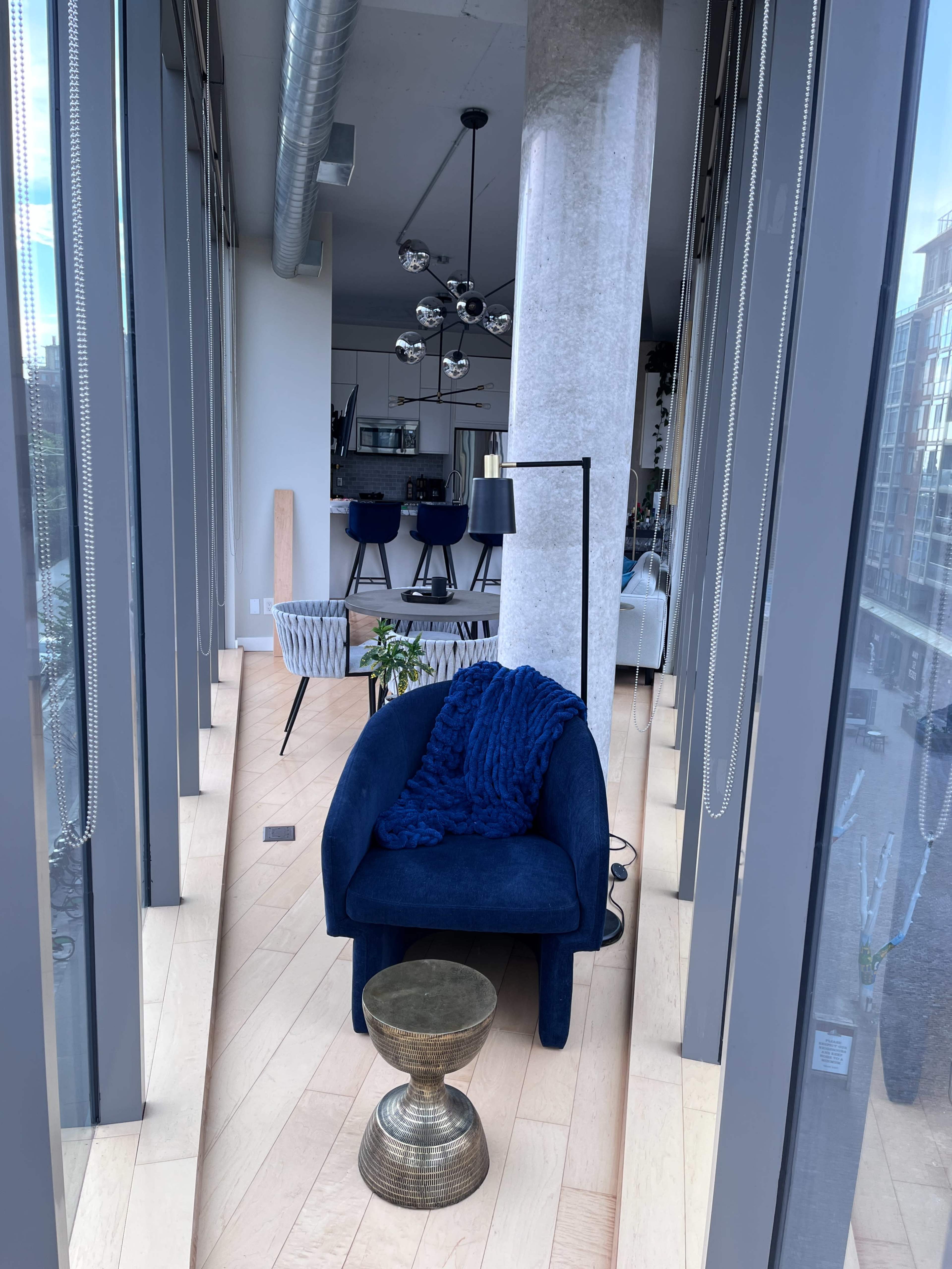 The image shows a modern interior space featuring a blue chair, a small metallic side table, and large windows that offer a view of the outside.