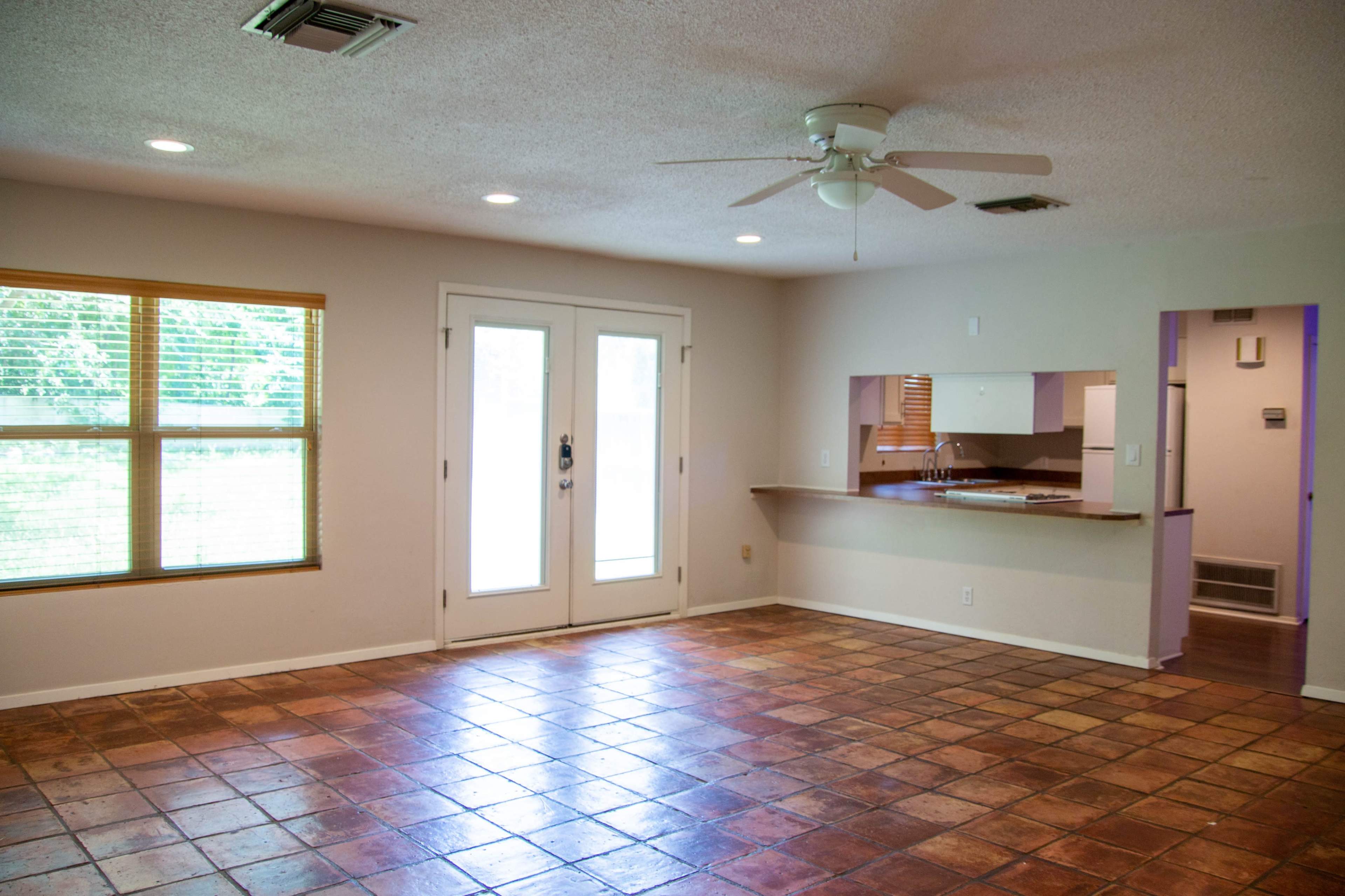 The image shows a spacious living room with tile flooring, a ceiling fan, and a set of double doors leading to an outdoor area.