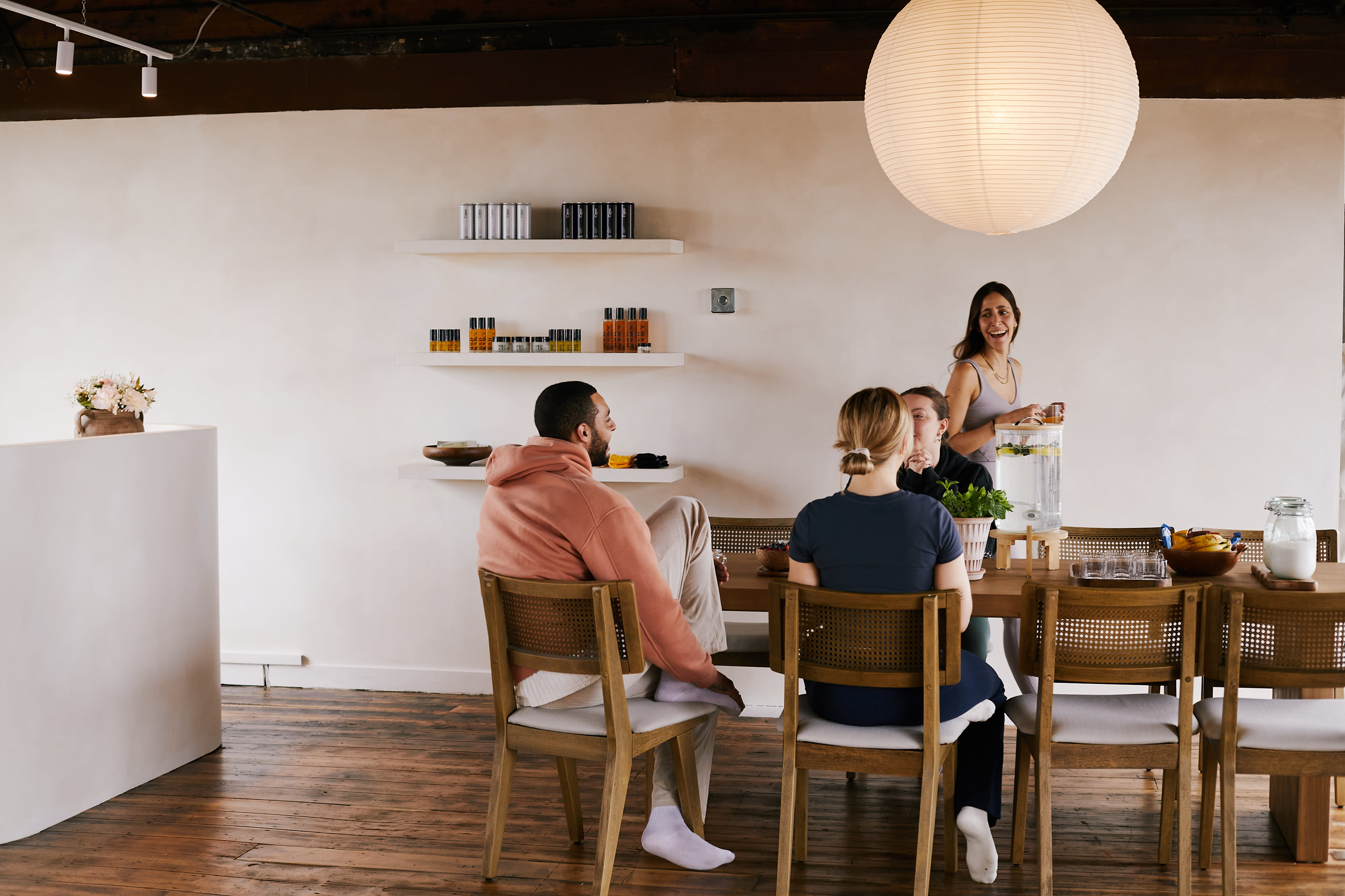 A group of four people sits around a wooden dining table in a bright, minimalist room with a large pendant light overhead.