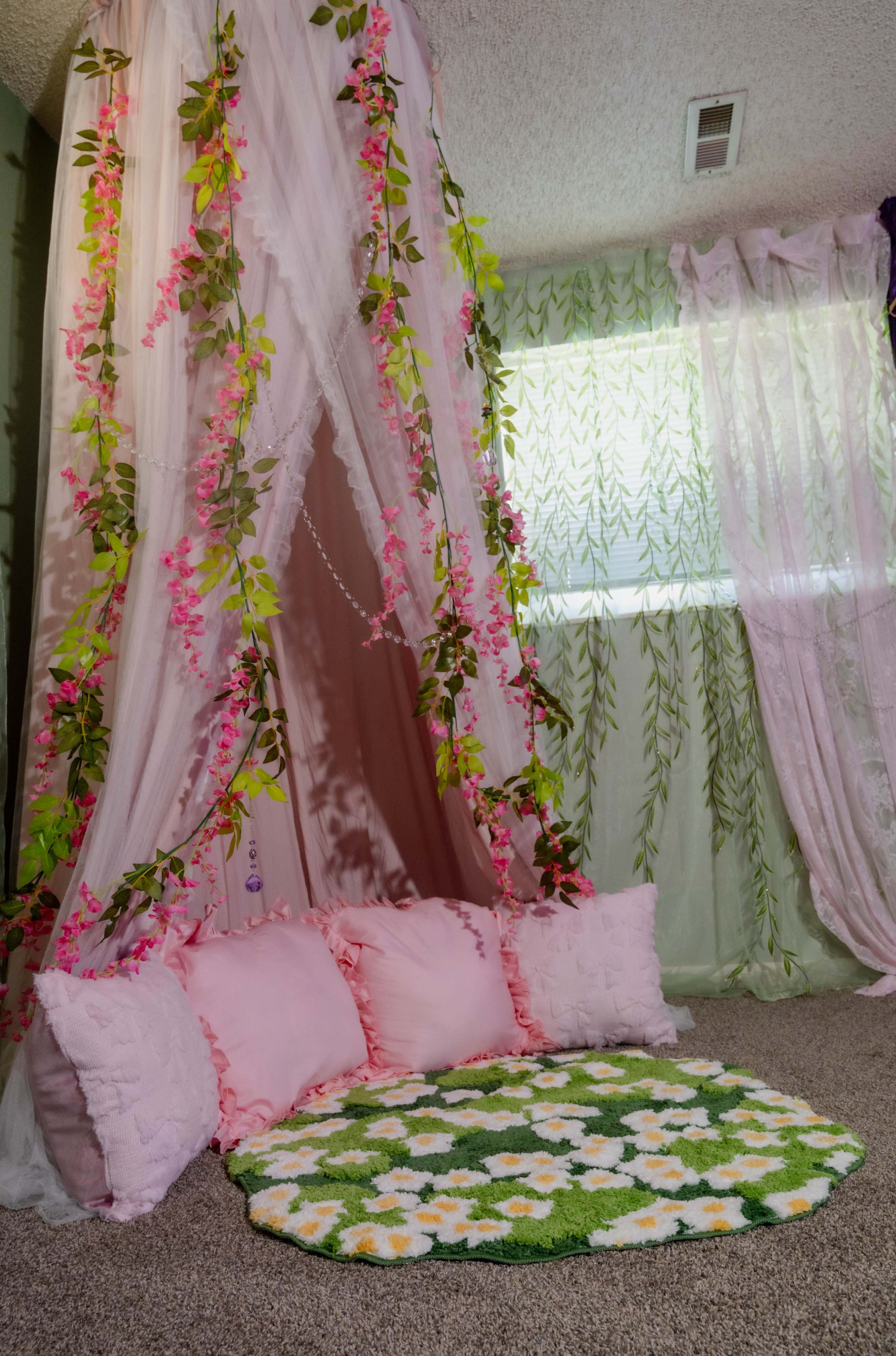 A cozy corner features a floral-themed rug, soft pink cushions, and sheer curtains draped with greenery and flowers.